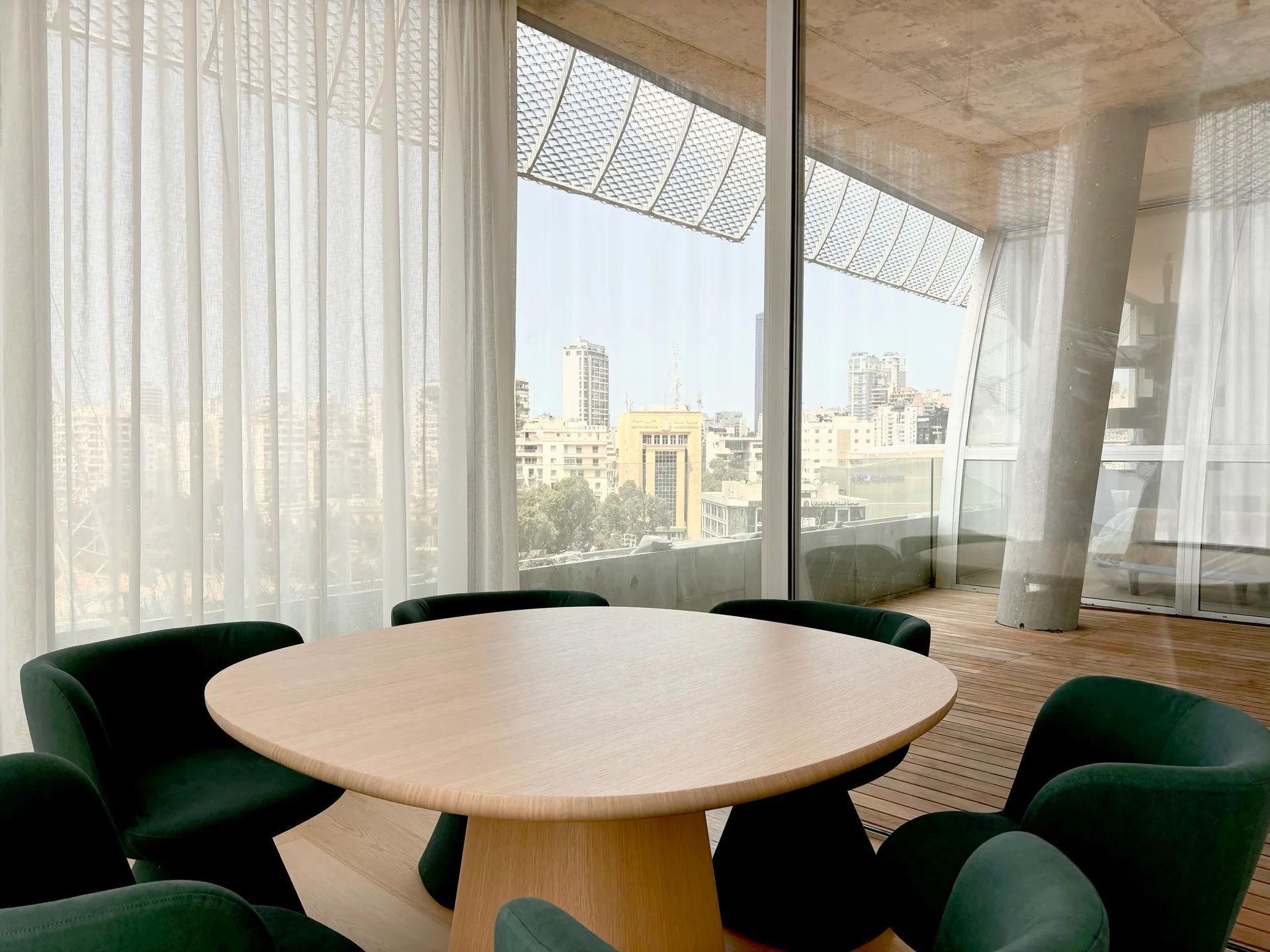 A round light wood dining table is surrounded by dark green upholstered chairs near a floor-to-ceiling window with sheer white curtains.