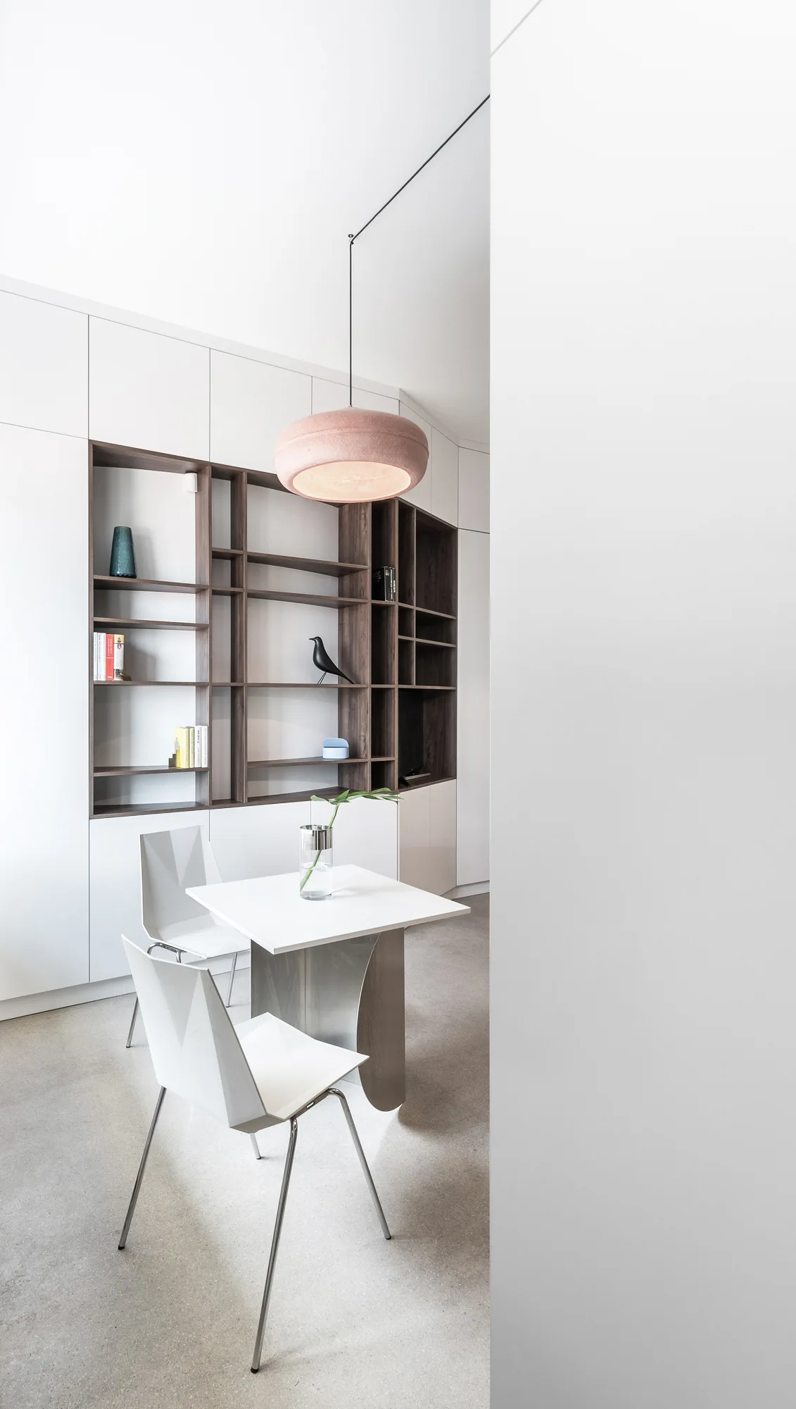 A minimalist dining area featuring a white table and angular chairs set against seamless white cabinets with a dark wood recessed shelving unit.