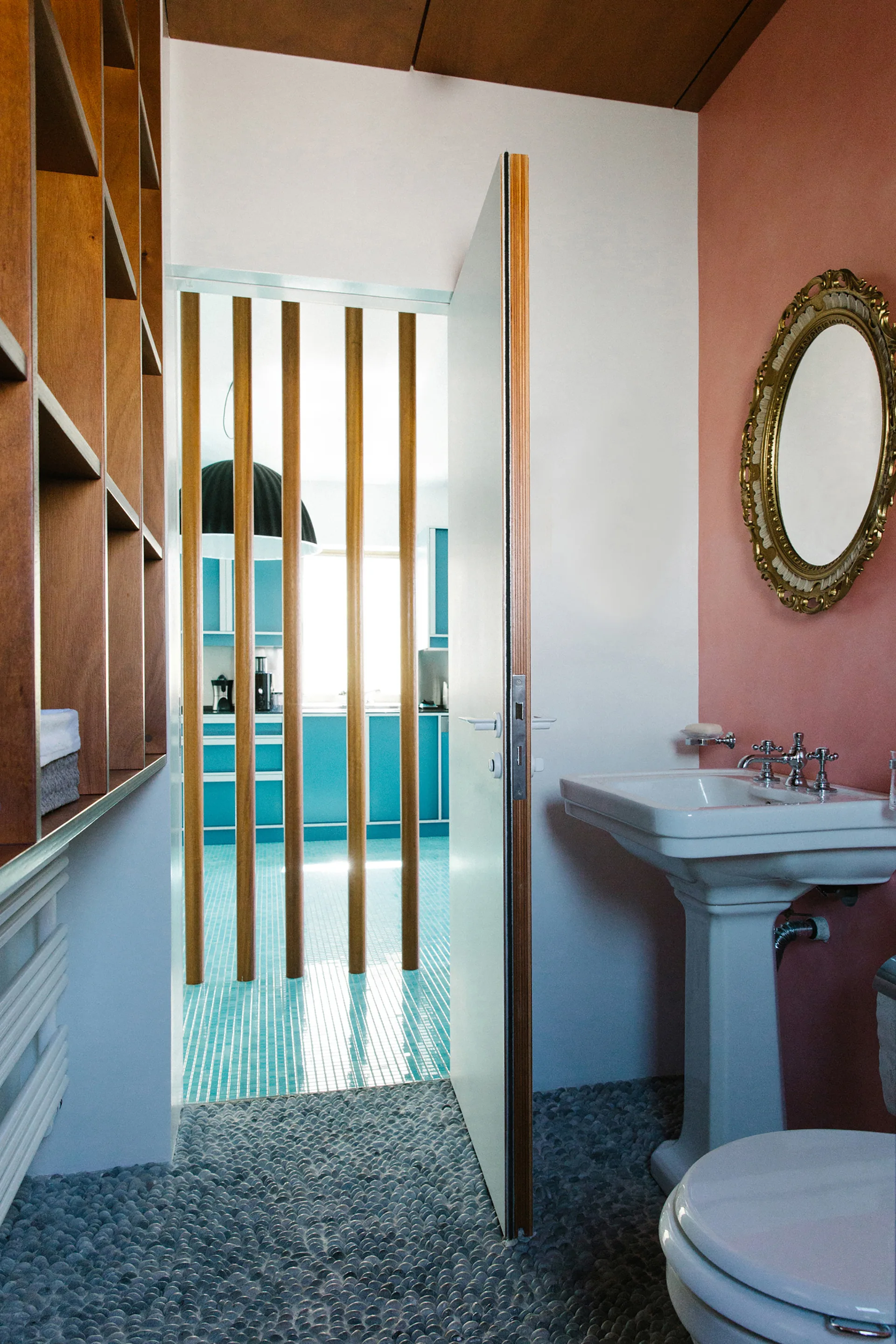 A bathroom features a pink wall, pedestal sink, and gray pebble floor, looking through a wood-slat divider into a turquoise kitchen.
