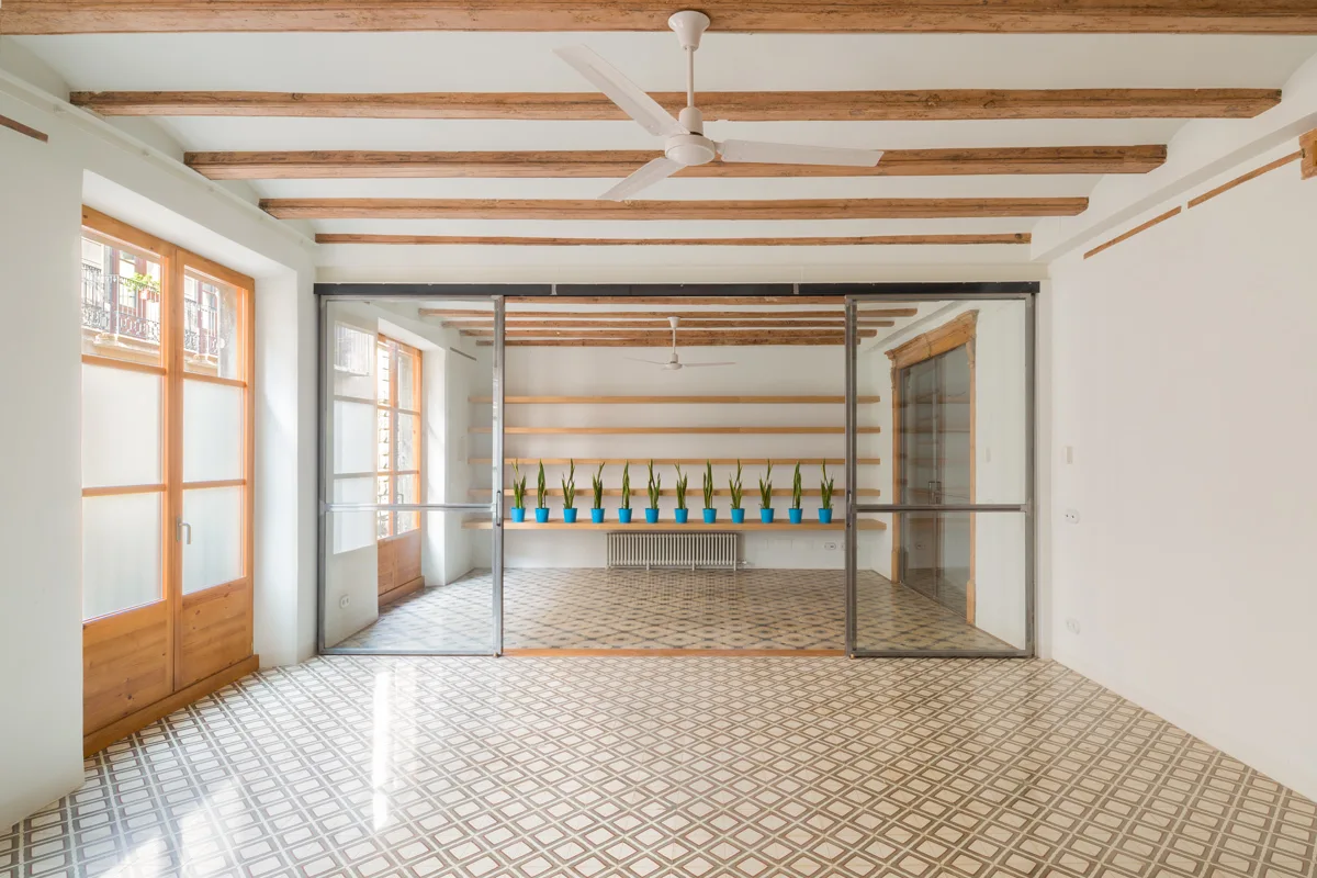 A minimalist room features patterned geometric tile flooring, exposed wood ceiling beams, and a sliding glass partition.