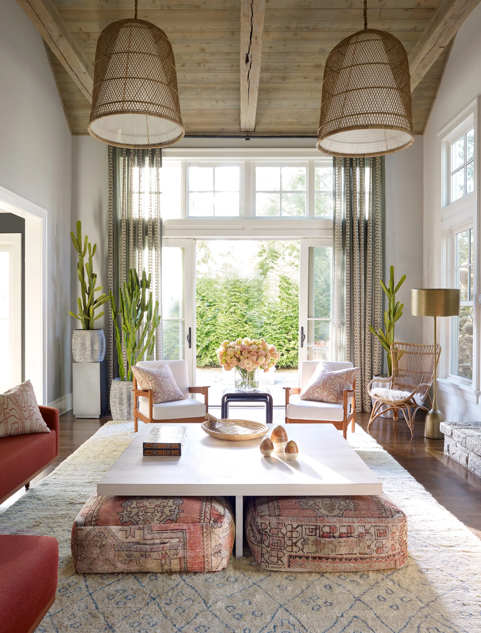A bright living room features a vaulted wood ceiling, large woven pendant lights, two armchairs, and patterned floor poufs beneath a coffee table.