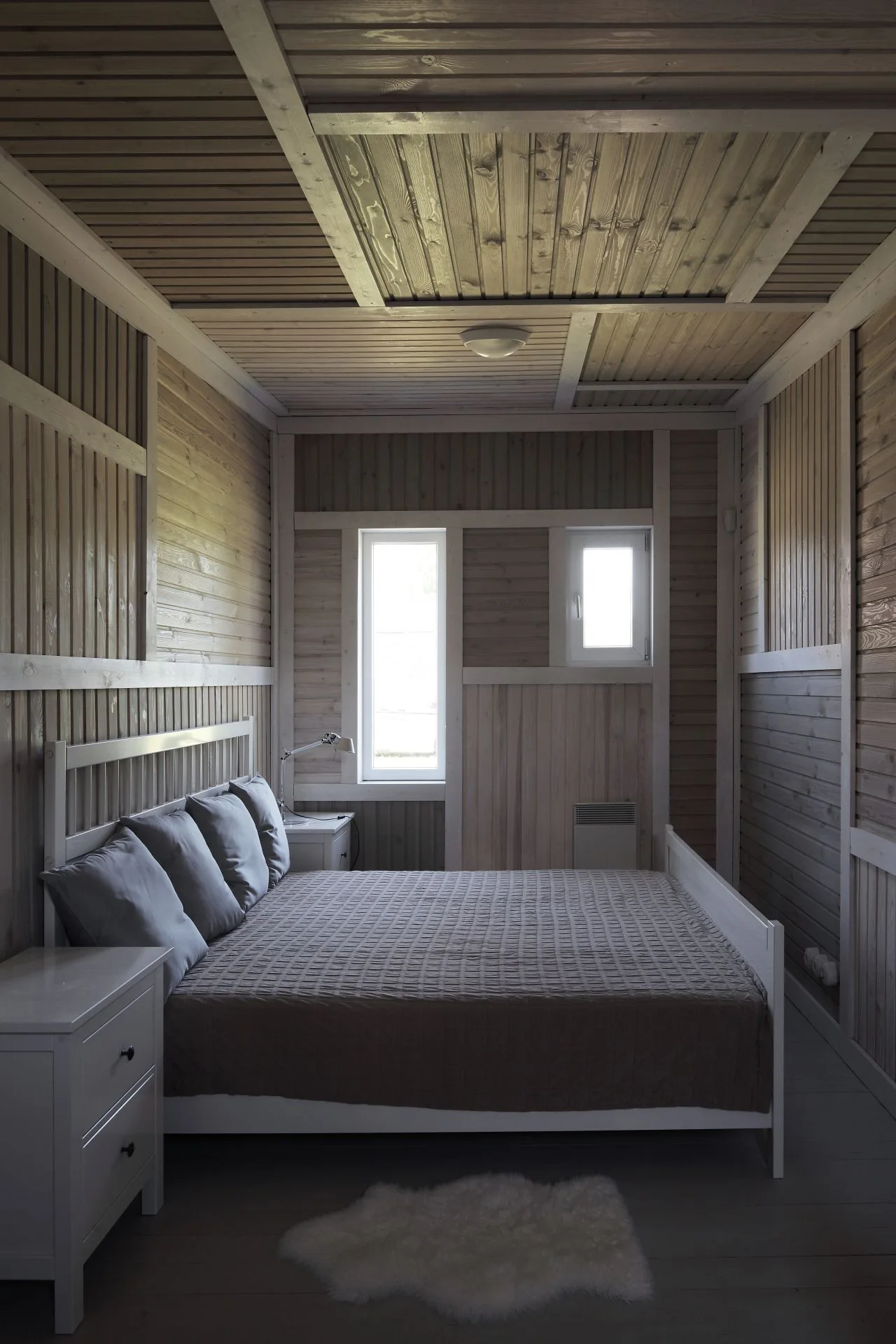 A rustic bedroom features a white bed frame, grey pillows, and matching white nightstands set against light wood-paneled walls and ceiling.