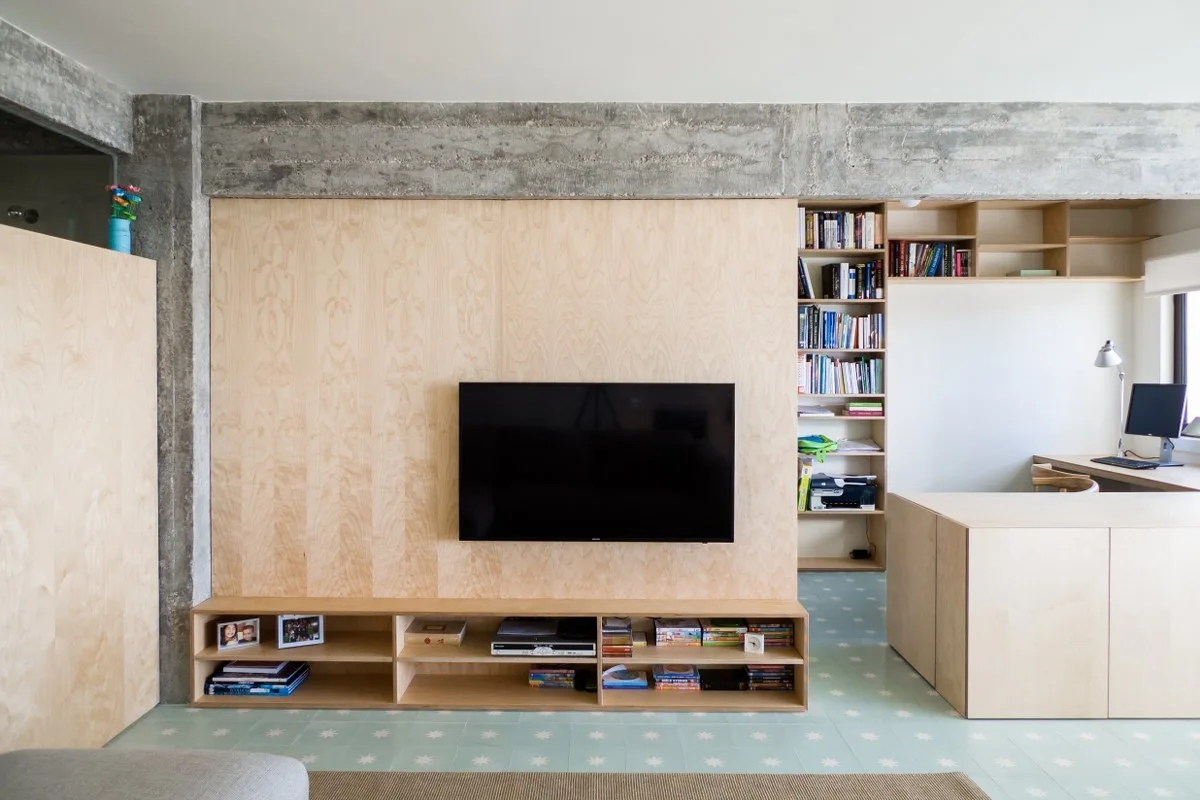 A living room featuring a plywood media wall, an exposed concrete beam, matching bookshelves, and light blue star-patterned floor tiles.
