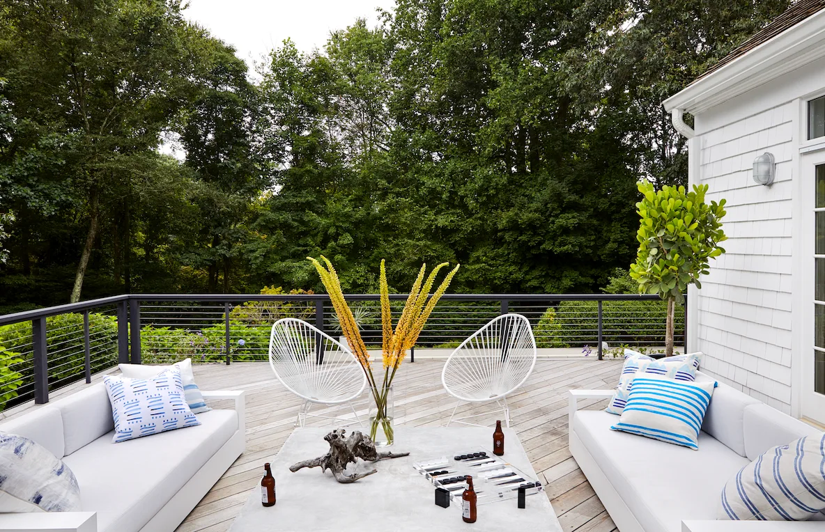 A modern wood-planked outdoor deck featuring white sofas, corded chairs, and a large concrete coffee table against a backdrop of dense green trees.