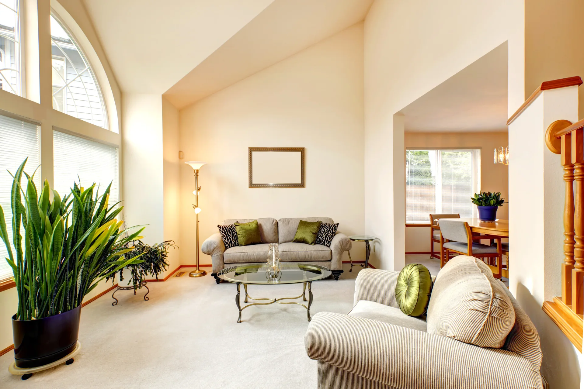A bright living room featuring a vaulted ceiling, a large arched window, beige sofas, a glass coffee table, and green accent pillows.