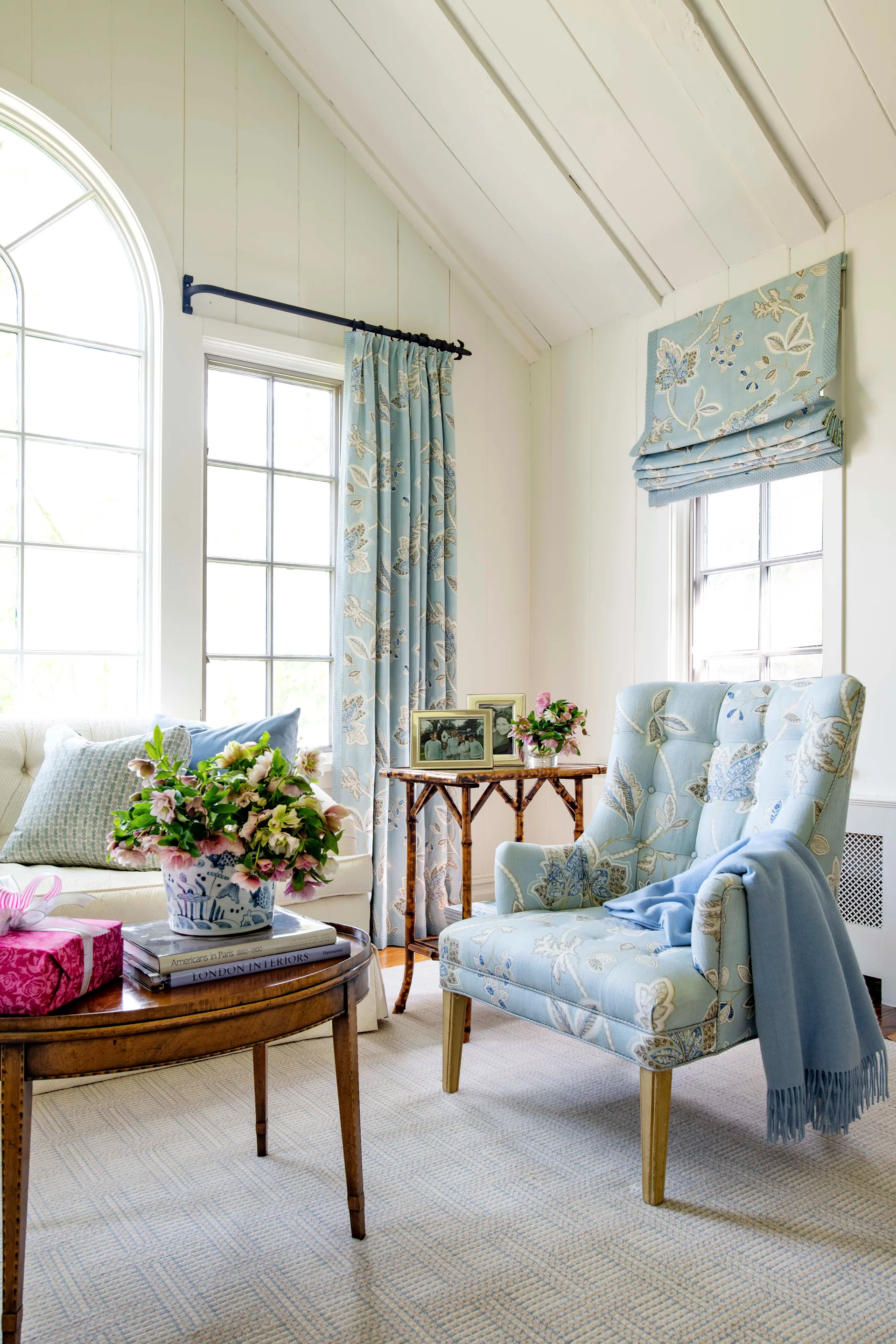 A bright living room corner features a blue floral armchair, matching patterned curtains, and a bamboo side table next to a white wood-paneled wall.