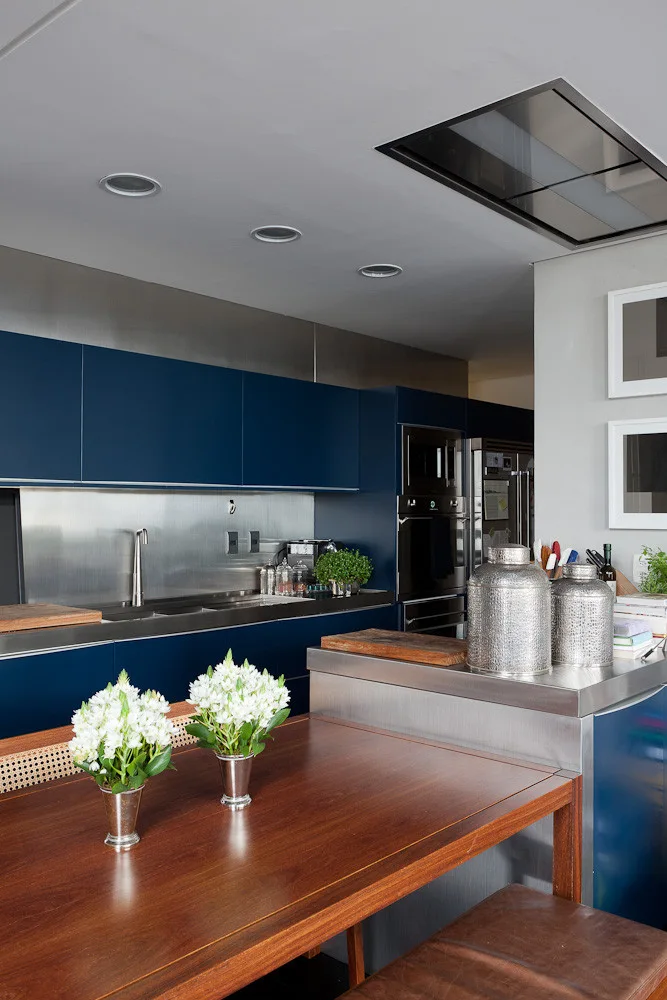A modern kitchen featuring matte navy blue cabinets, a stainless steel backsplash and countertop, and a wooden dining table in the foreground.