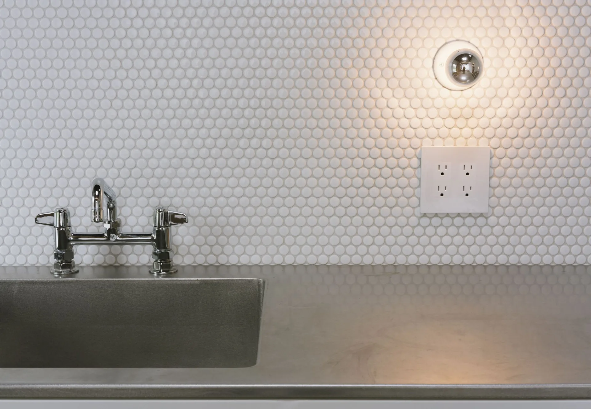 A chrome faucet and integrated steel sink are set against a white penny tile backsplash with a glowing spherical sconce.