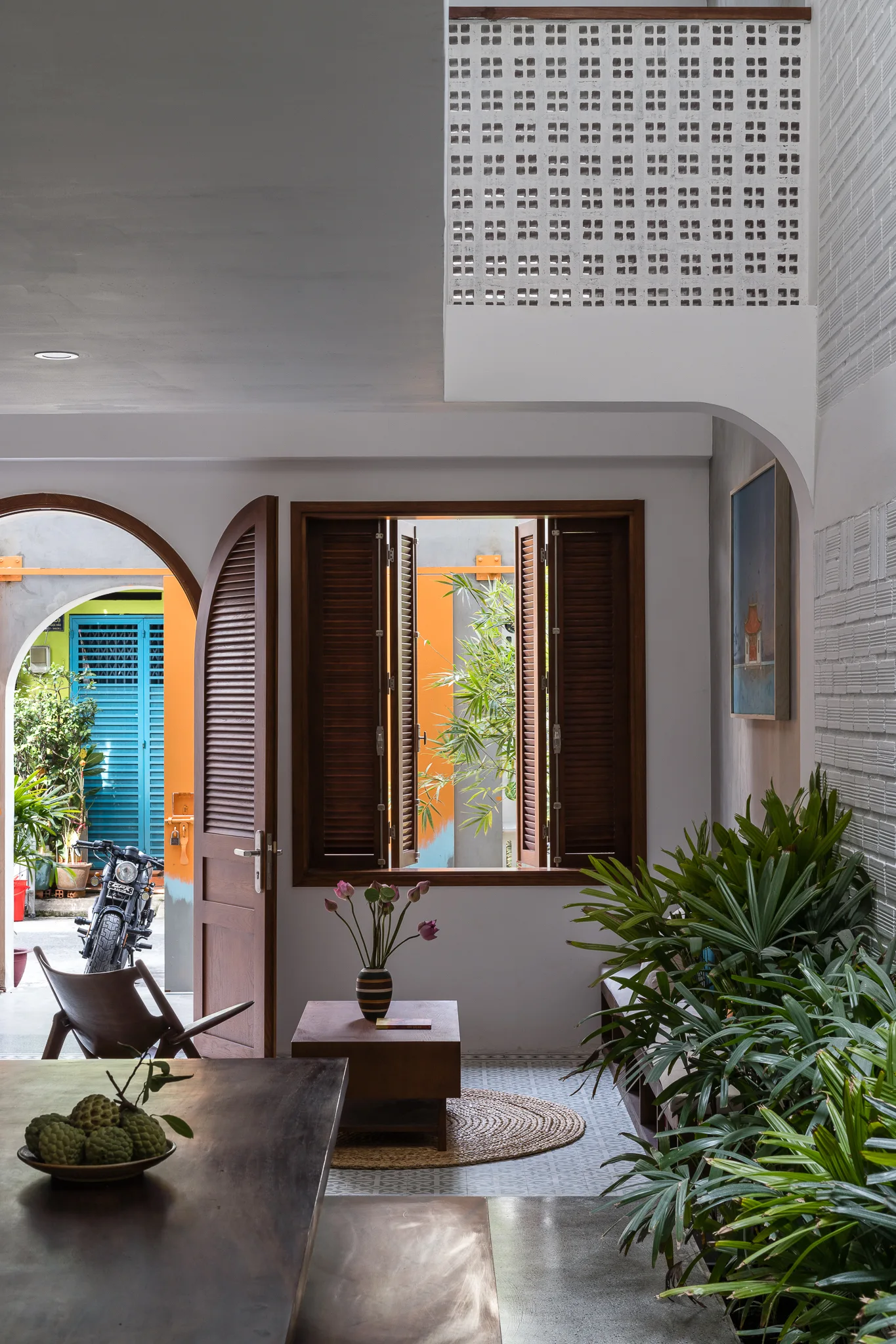 A tropical modern living area features an arched entryway, wooden louvered shutters, patterned floor tiles, and a collection of lush green potted plants.