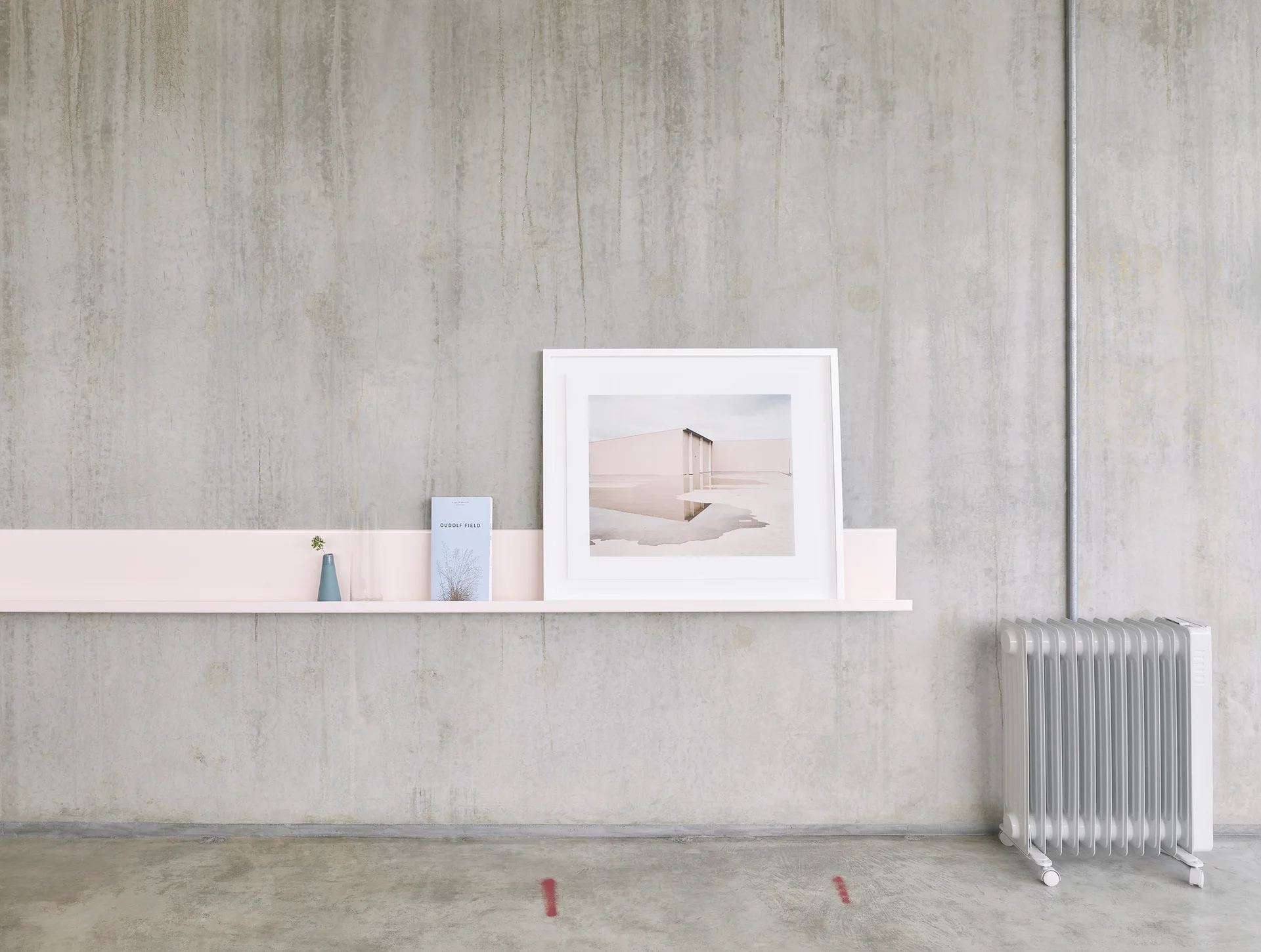 A long pale pink wall shelf holding a framed photo print, a book, and a small vase is mounted on a textured concrete wall next to a white radiator.