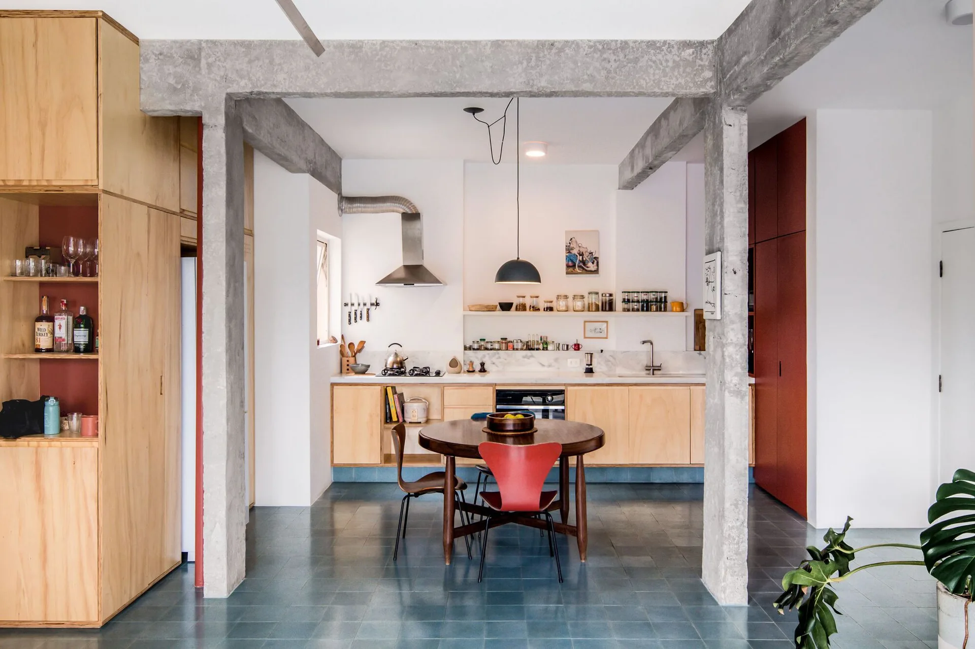 A modern kitchen and dining area features light plywood cabinetry, exposed concrete structural beams, a round wooden table, and blue floor tiles.