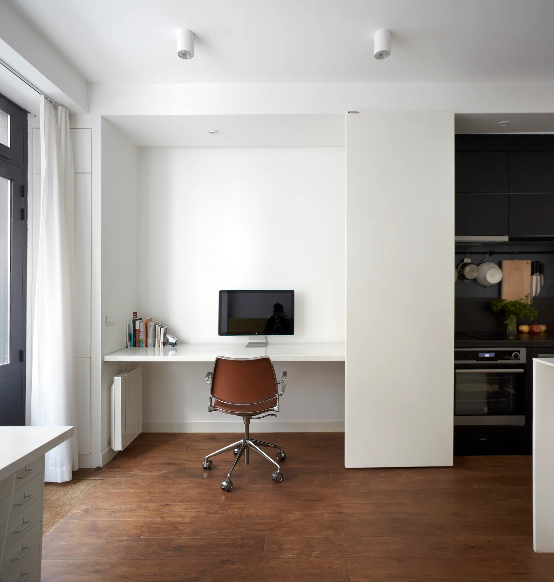 A minimalist home office features a white floating desk with a monitor, a brown swivel chair, wood floors, and a sliding white partition.