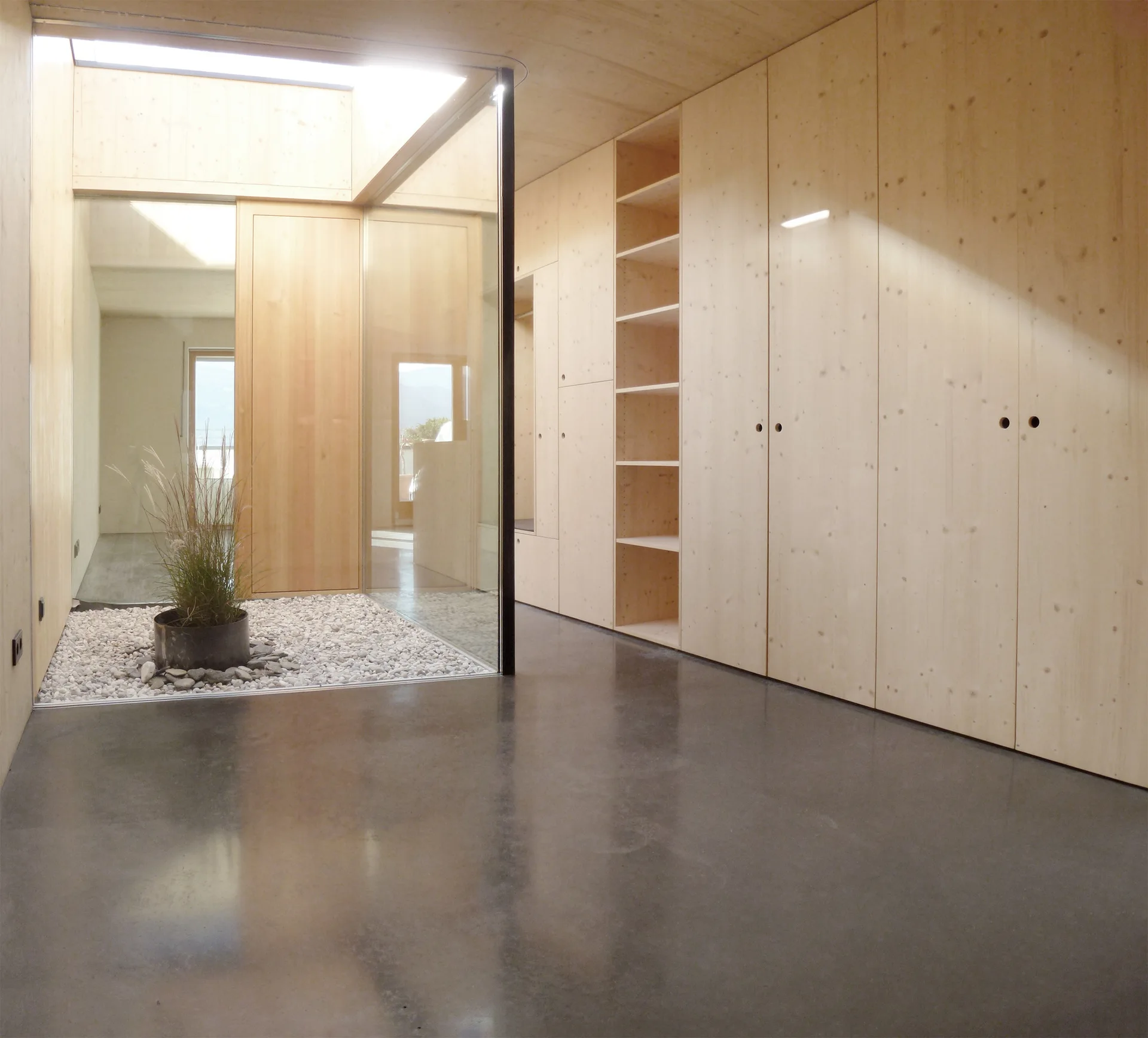 A minimalist hallway features a polished concrete floor, a wall of light wood built-in cabinets, and a glass-enclosed indoor courtyard with white pebbles.