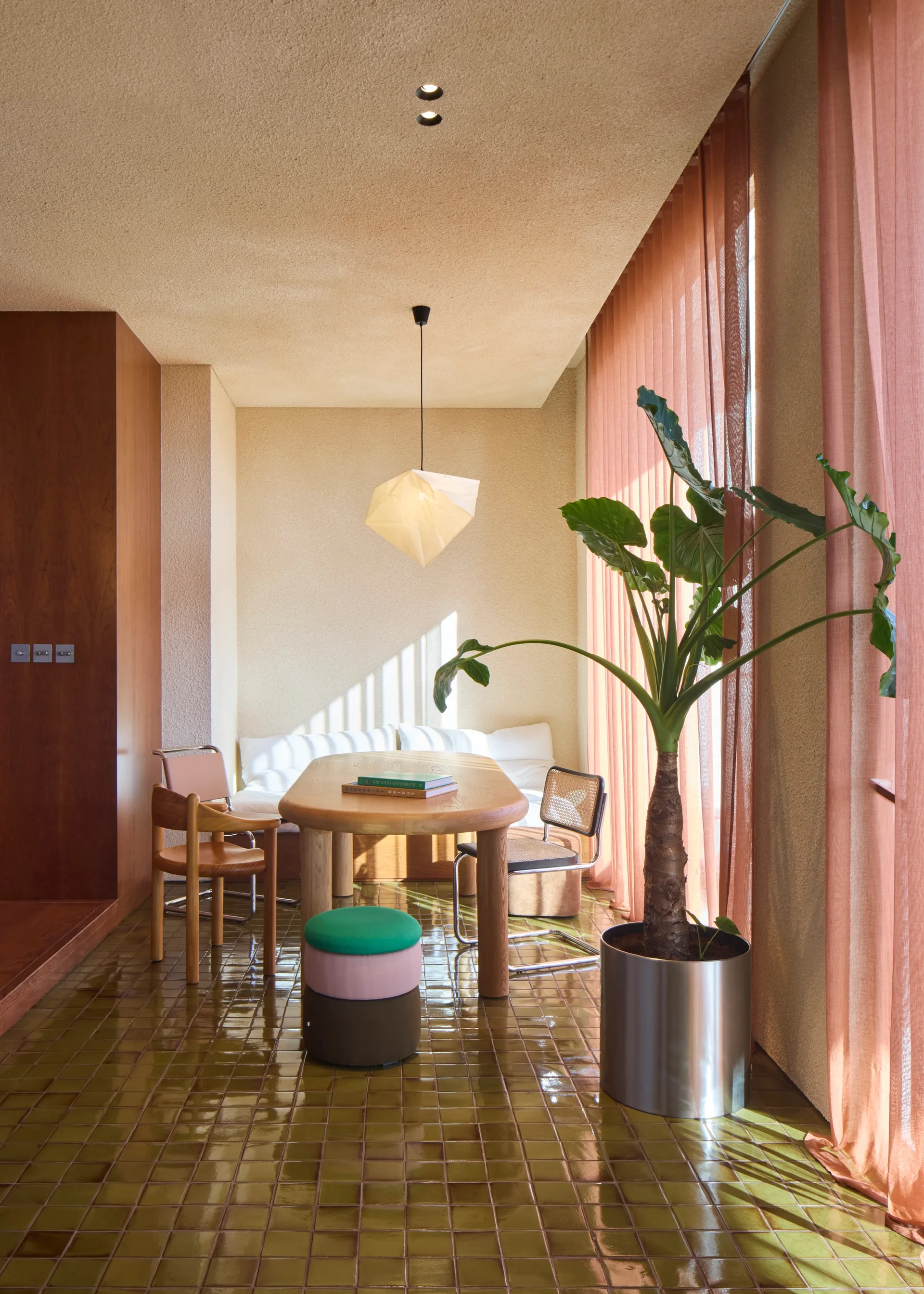 An oval wooden dining table sits on a glossy olive green tile floor near textured beige plaster walls and sheer terracotta curtains.
