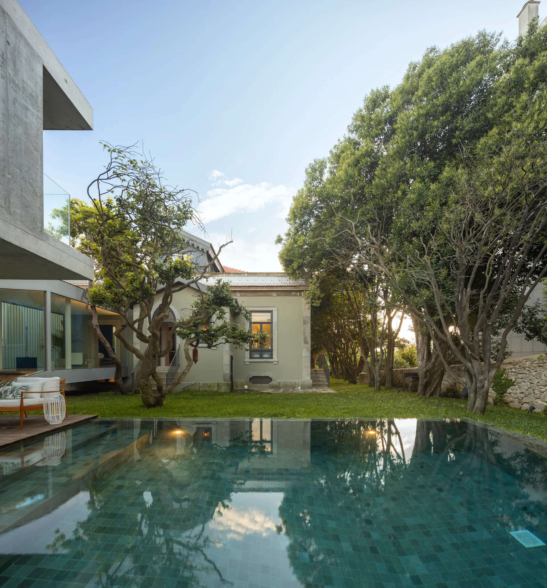 A dark teal tiled swimming pool sits next to a green lawn, mature trees, and a modern concrete residential extension.