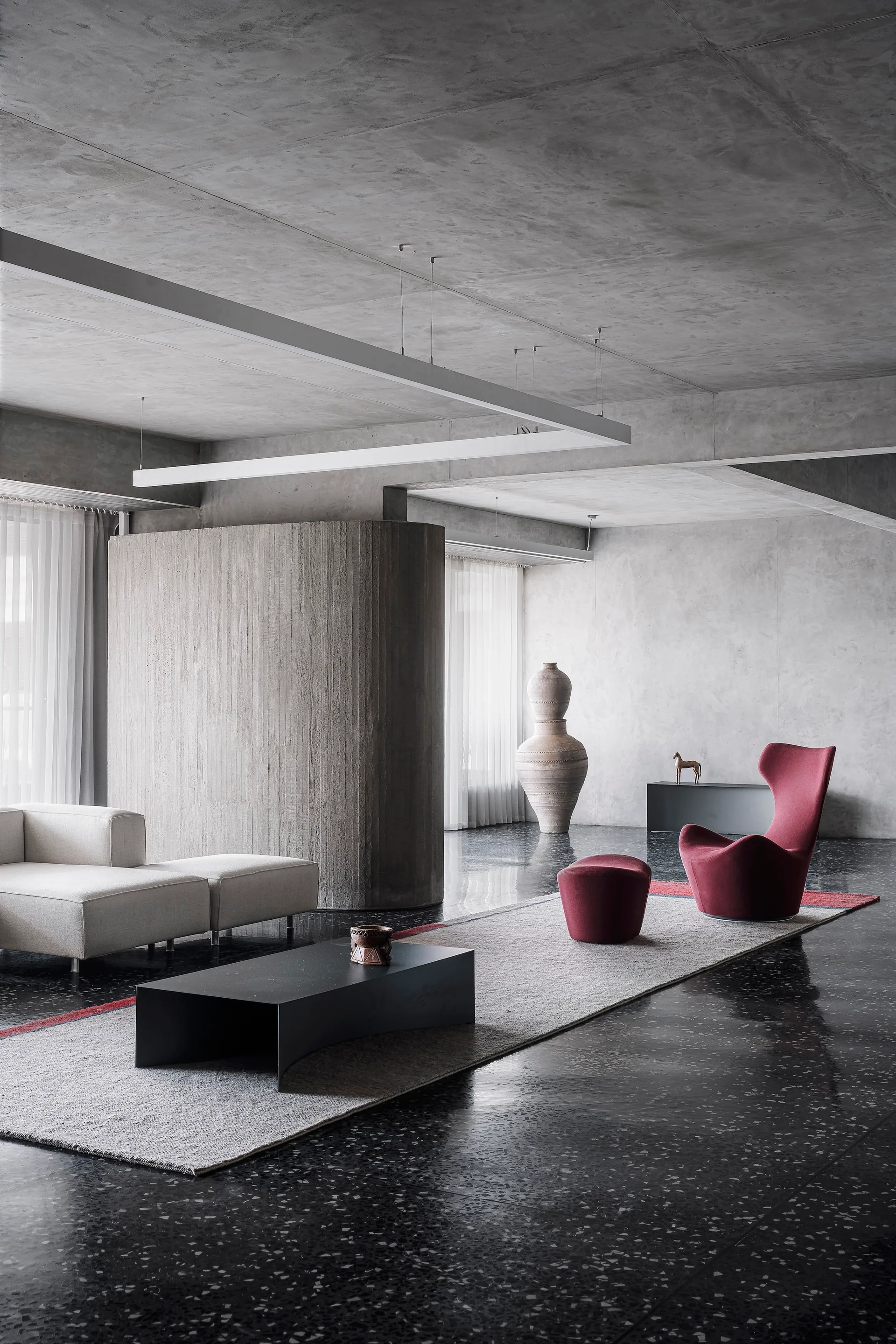 A minimalist brutalist living room features a curved concrete partition wall, a cream sectional sofa, a black coffee table, and a sculptural red armchair.