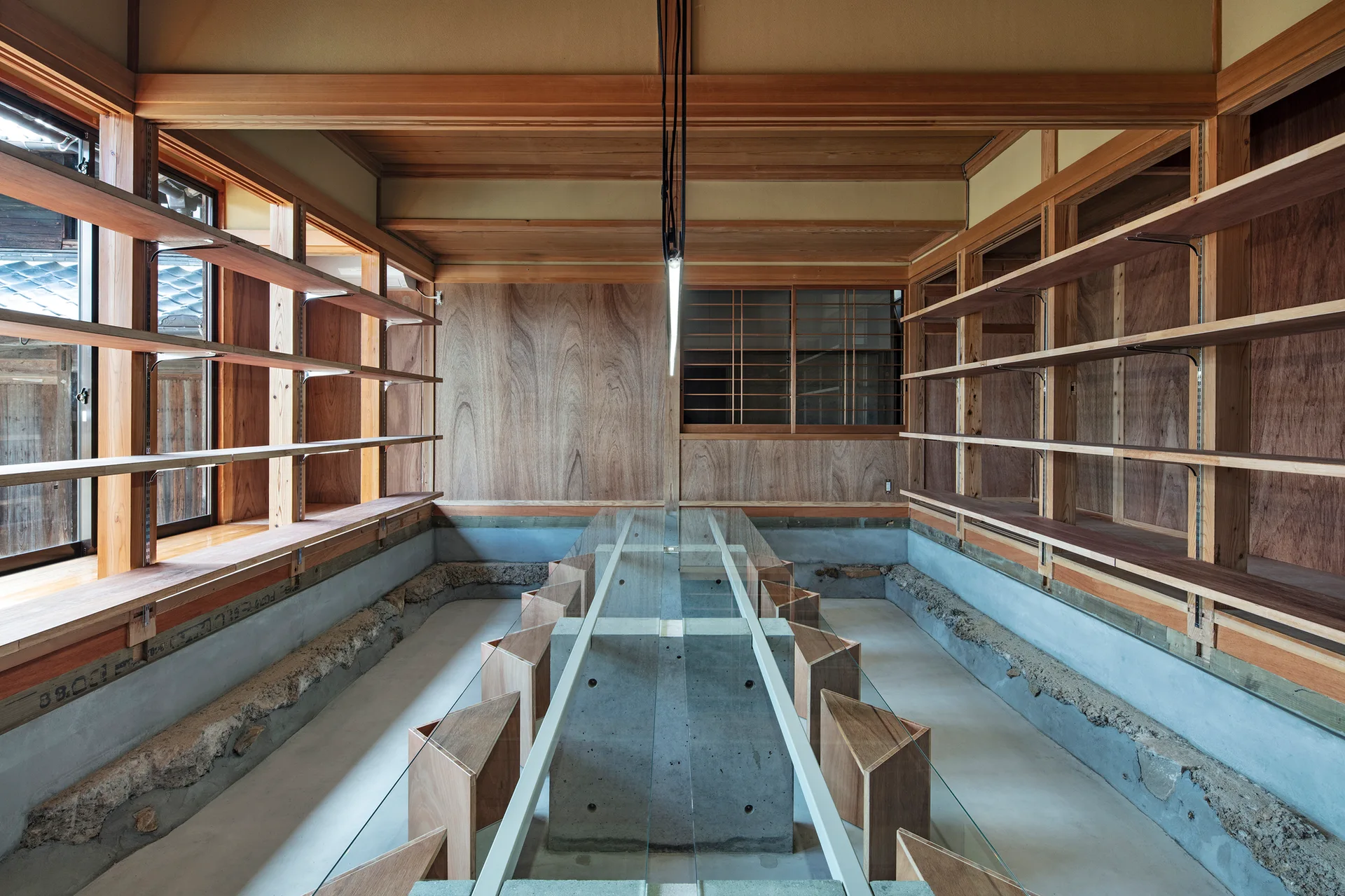A minimalist room features a long concrete and glass table lined with triangular wooden stools and flanked by open wooden shelving units.