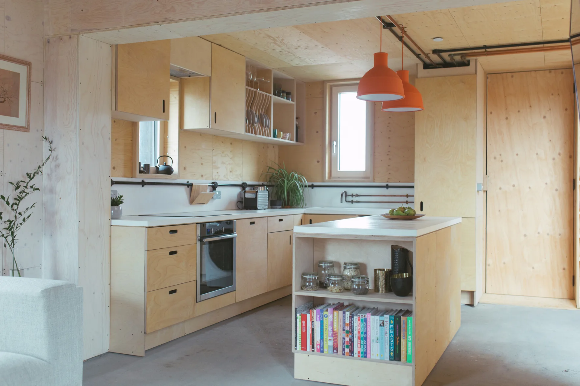 A minimalist plywood kitchen features natural wood cabinetry, a central island with integrated book storage, and two orange pendant lights.
