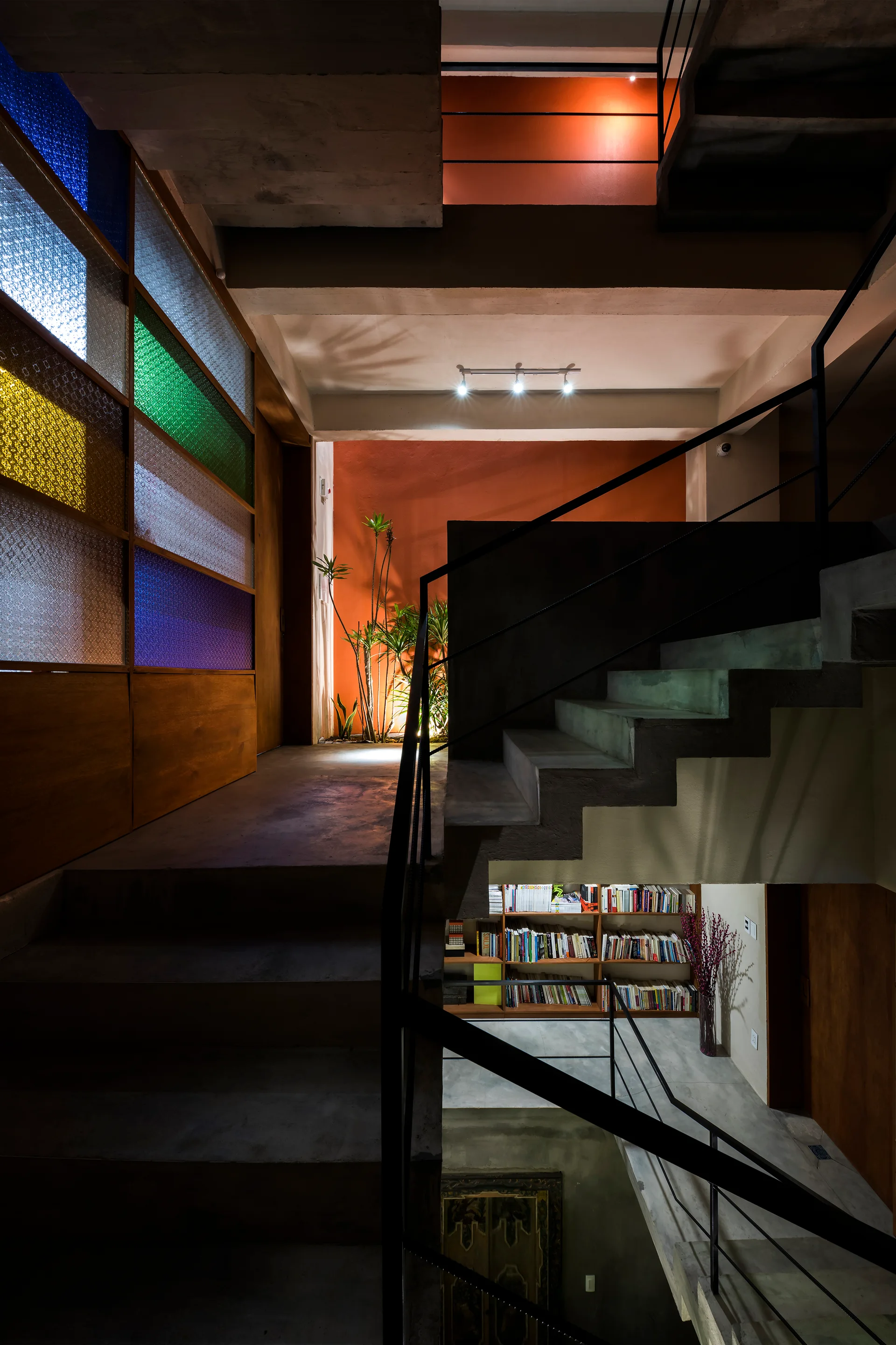 A multi-level concrete staircase features black metal railings, a bold orange accent wall, and a partition made of colorful textured glass panels.