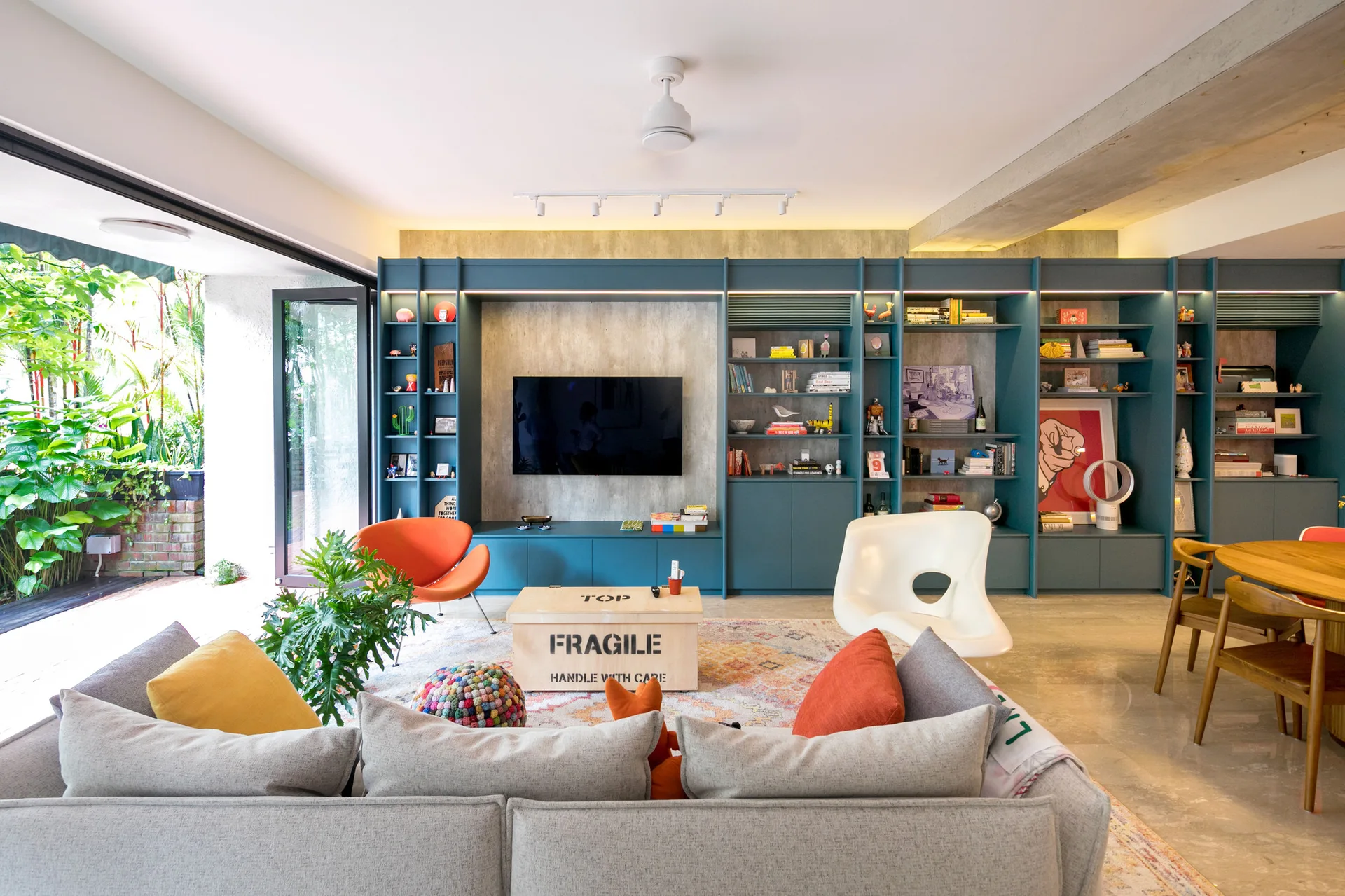 A modern living room features a large blue built-in shelving unit, a grey sectional sofa, an orange lounge chair, and a wooden crate coffee table.