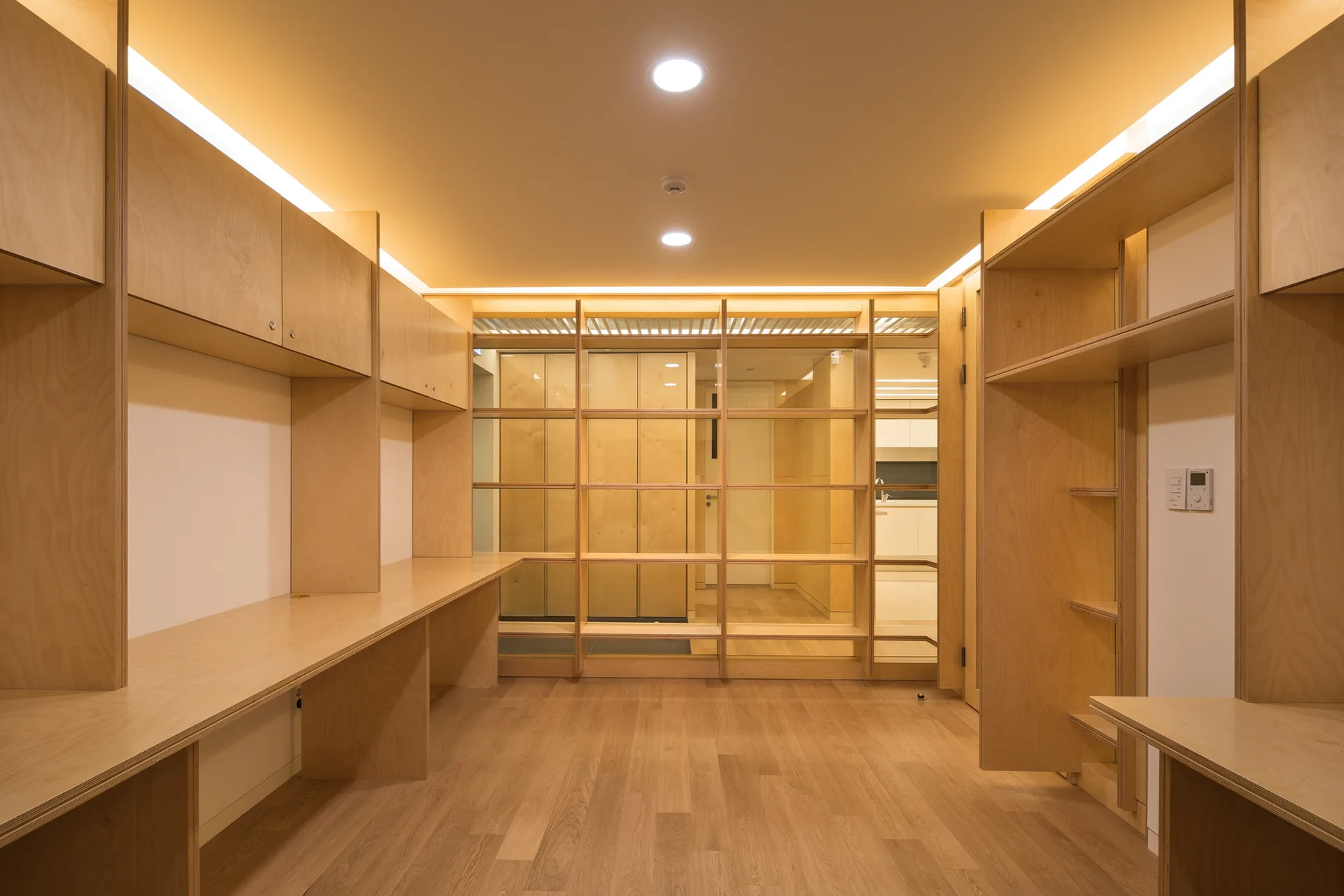 A minimalist home office features custom light plywood built-in desks, matching wall cabinets, open shelving, and integrated ceiling cove lighting.
