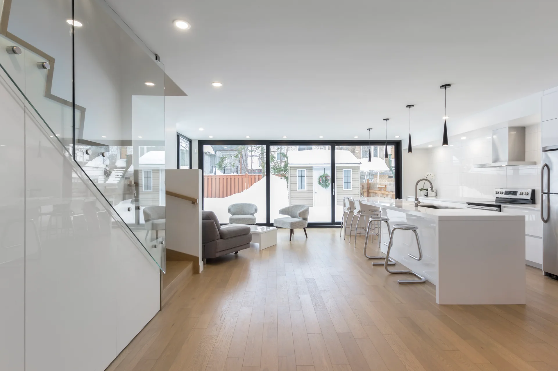 An open-concept kitchen and living area features a white kitchen island, black pendant lights, light wood flooring, and a staircase with a glass railing.