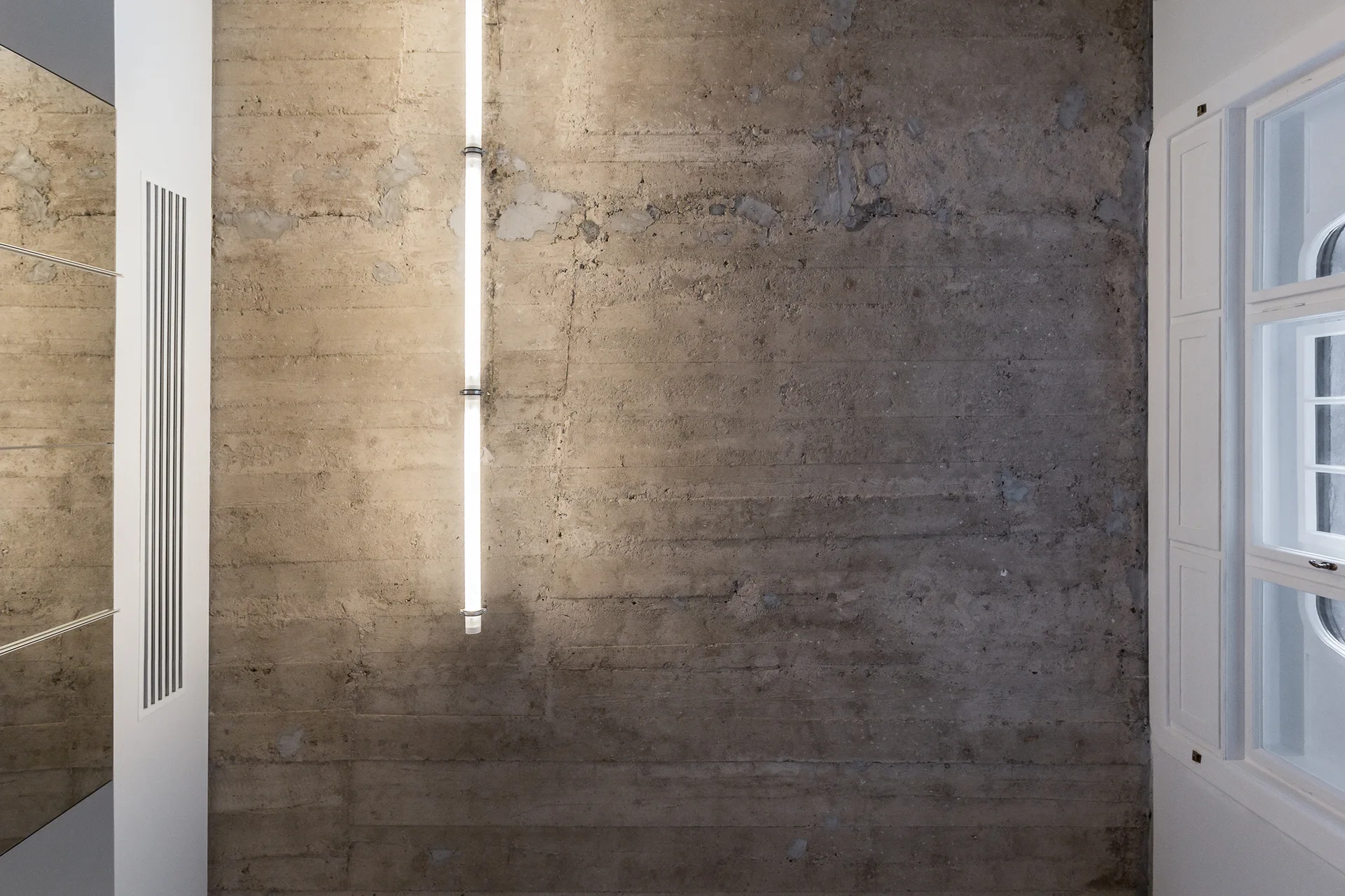 A raw textured concrete wall features a vertical tube light fixture alongside a white paneled window frame.