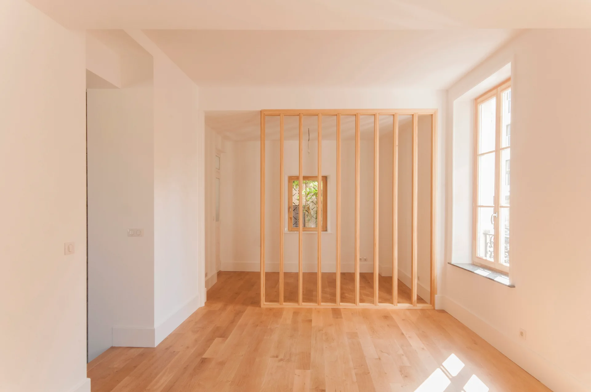 An empty white room features light hardwood flooring, a window, and a vertical wood slat partition in the center.