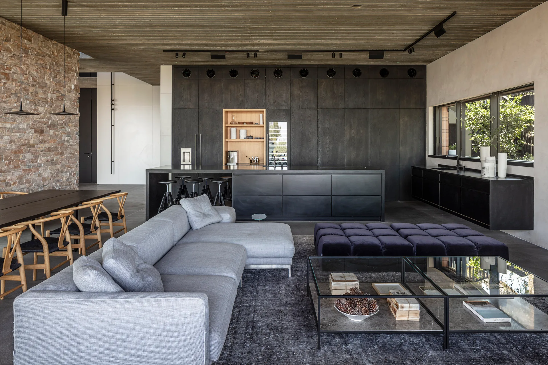 A spacious open-concept living area featuring a grey sectional sofa, glass coffee table, and flat-front black kitchen cabinets under a slatted wood ceiling.