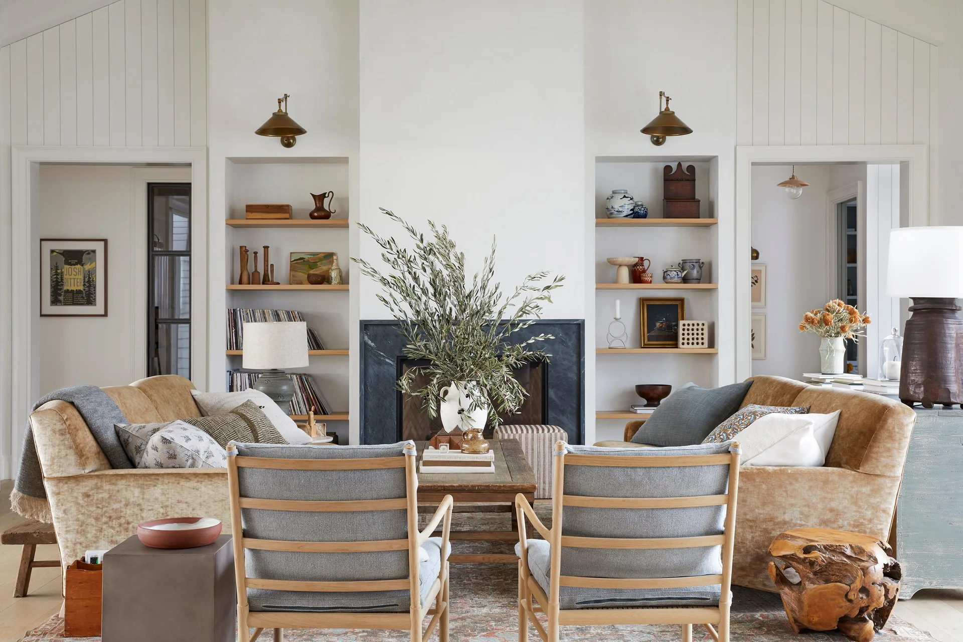 A symmetrical living room features a black stone fireplace flanked by built-in shelves, with tan velvet sofas and blue wooden armchairs.