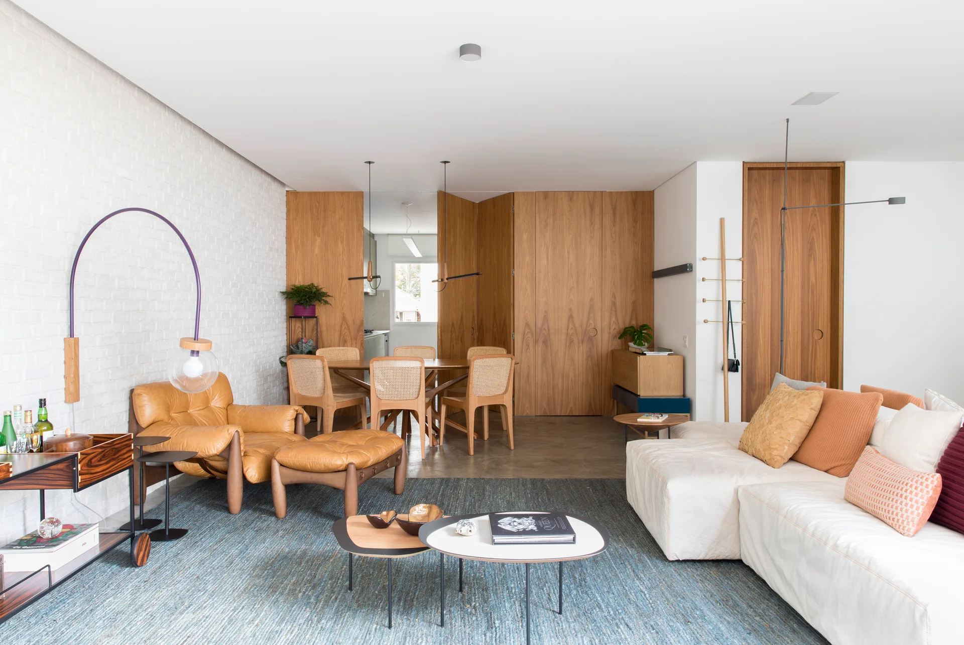 A modern open-concept living room with a white brick wall, tan leather armchair, and cream sofa on a blue rug in front of wood-paneled walls.