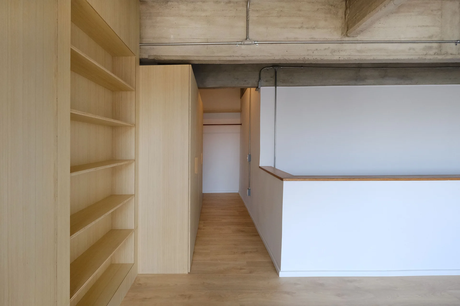 A minimalist hallway featuring custom blonde wood cabinetry, built-in shelving, a white half-wall, and an exposed raw concrete ceiling with metal conduits.