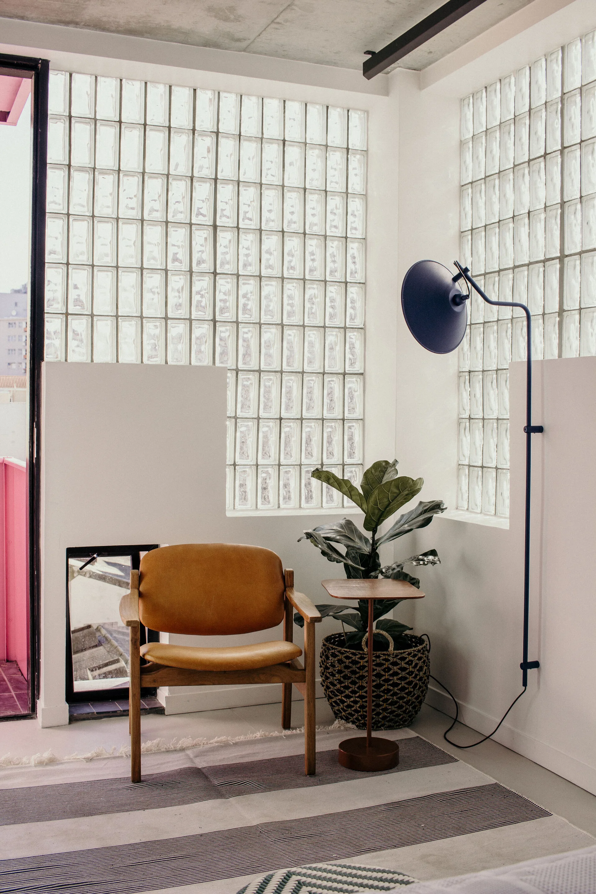 A tan leather armchair sits on a striped rug next to a fiddle leaf fig and a dark blue floor lamp against a glass block wall.