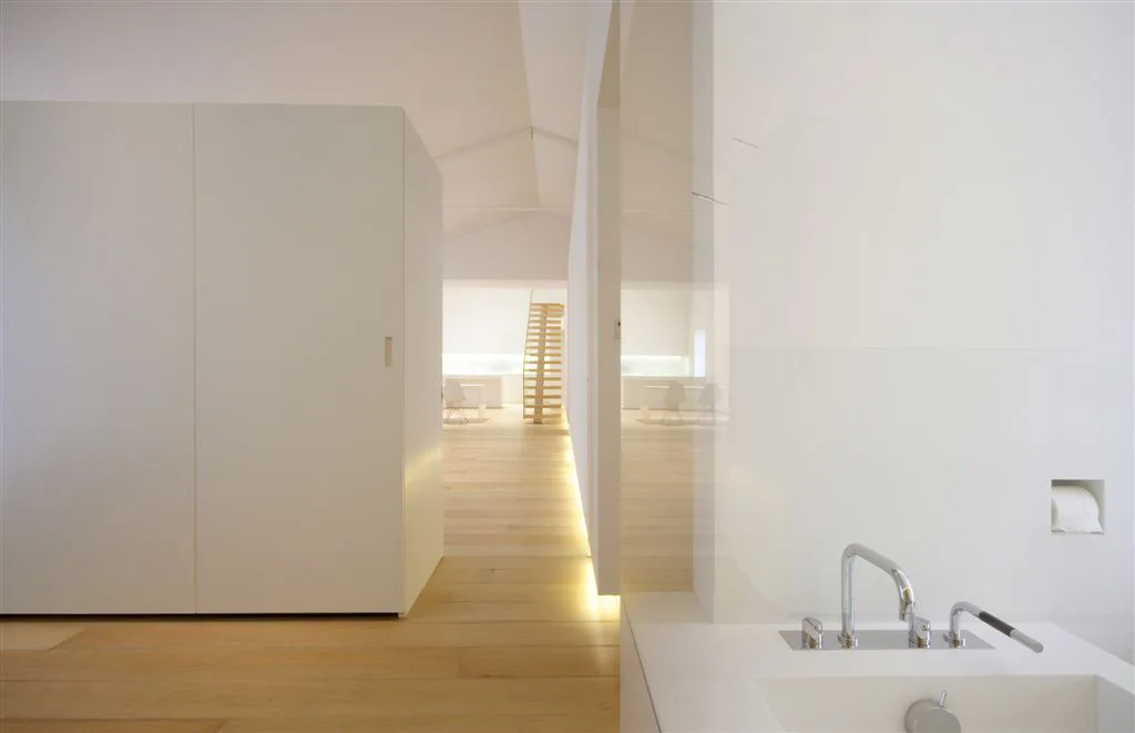 A minimalist white interior featuring light wood flooring, a modern chrome faucet, a white cabinet, and glowing baseboard strip lighting.