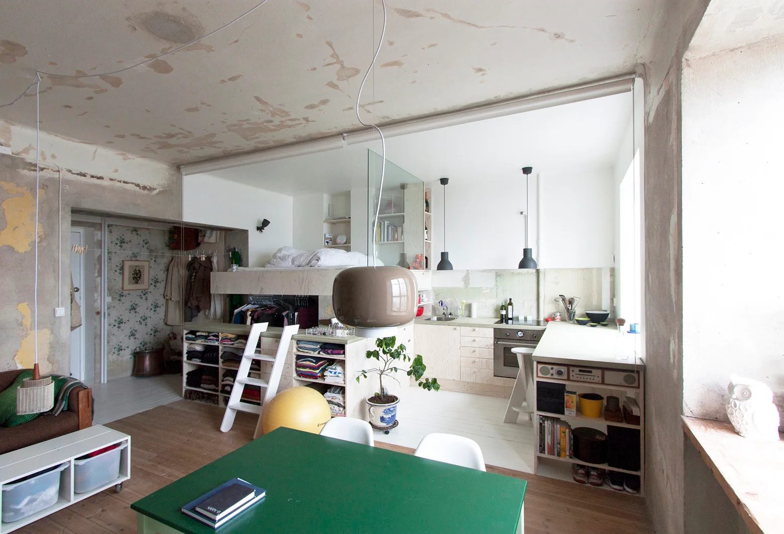 An industrial studio apartment features a lofted bed above a kitchen, a raw unfinished plaster ceiling, and a green dining table.