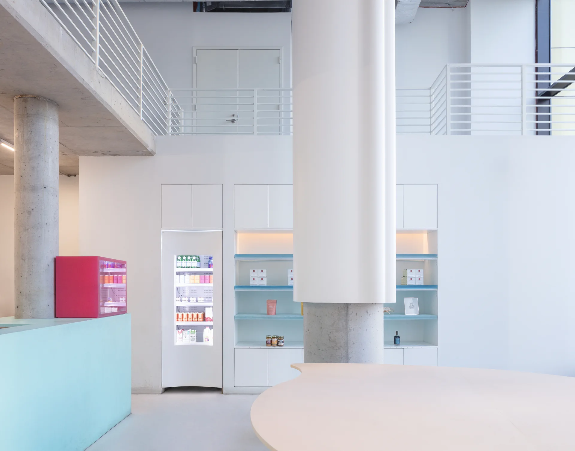 A minimalist retail interior features a large round column, built-in shelving with an integrated refrigerator, and a white mezzanine level.