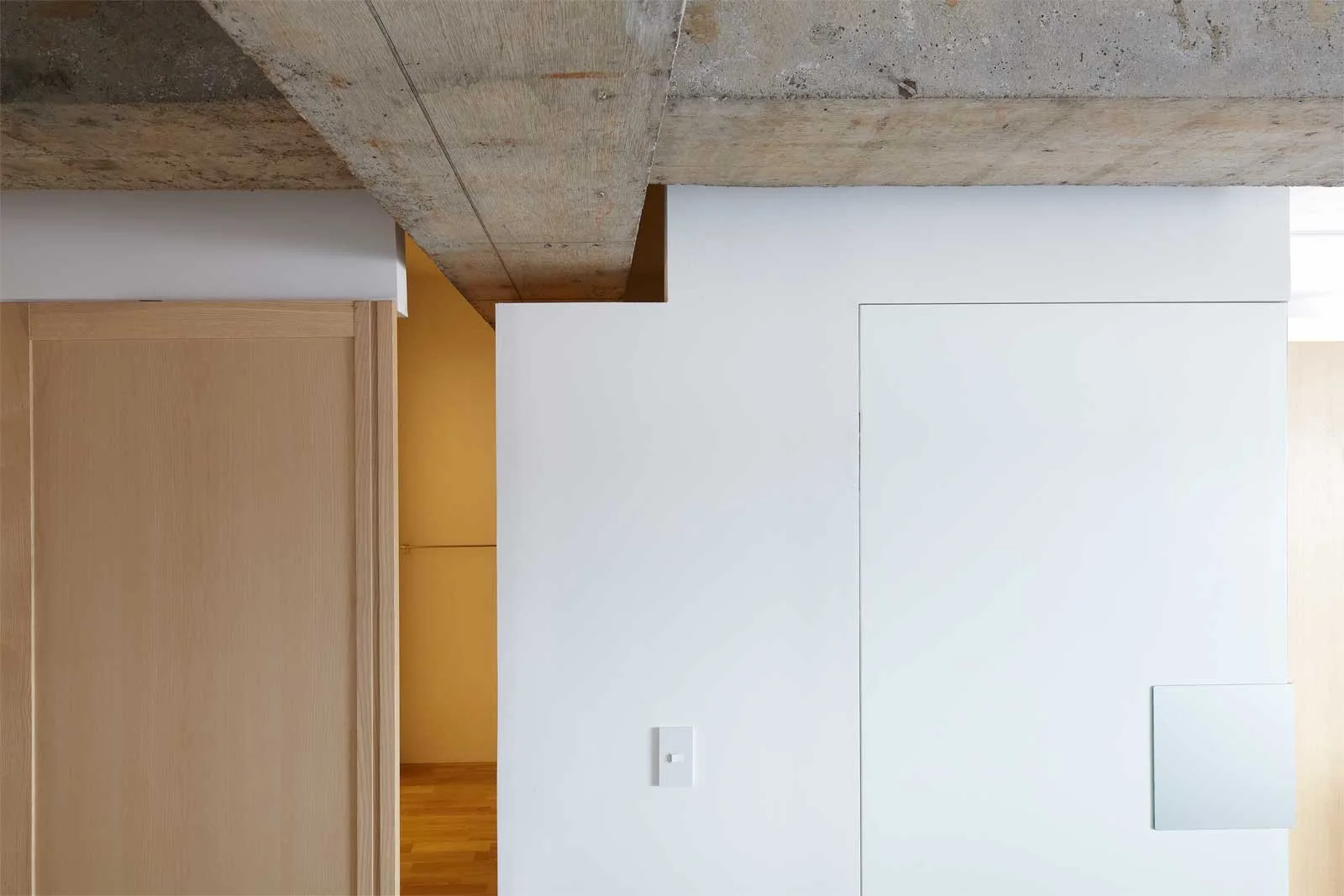 An exposed concrete ceiling beam runs above a smooth white partition wall and a light wood door frame in a minimalist interior.