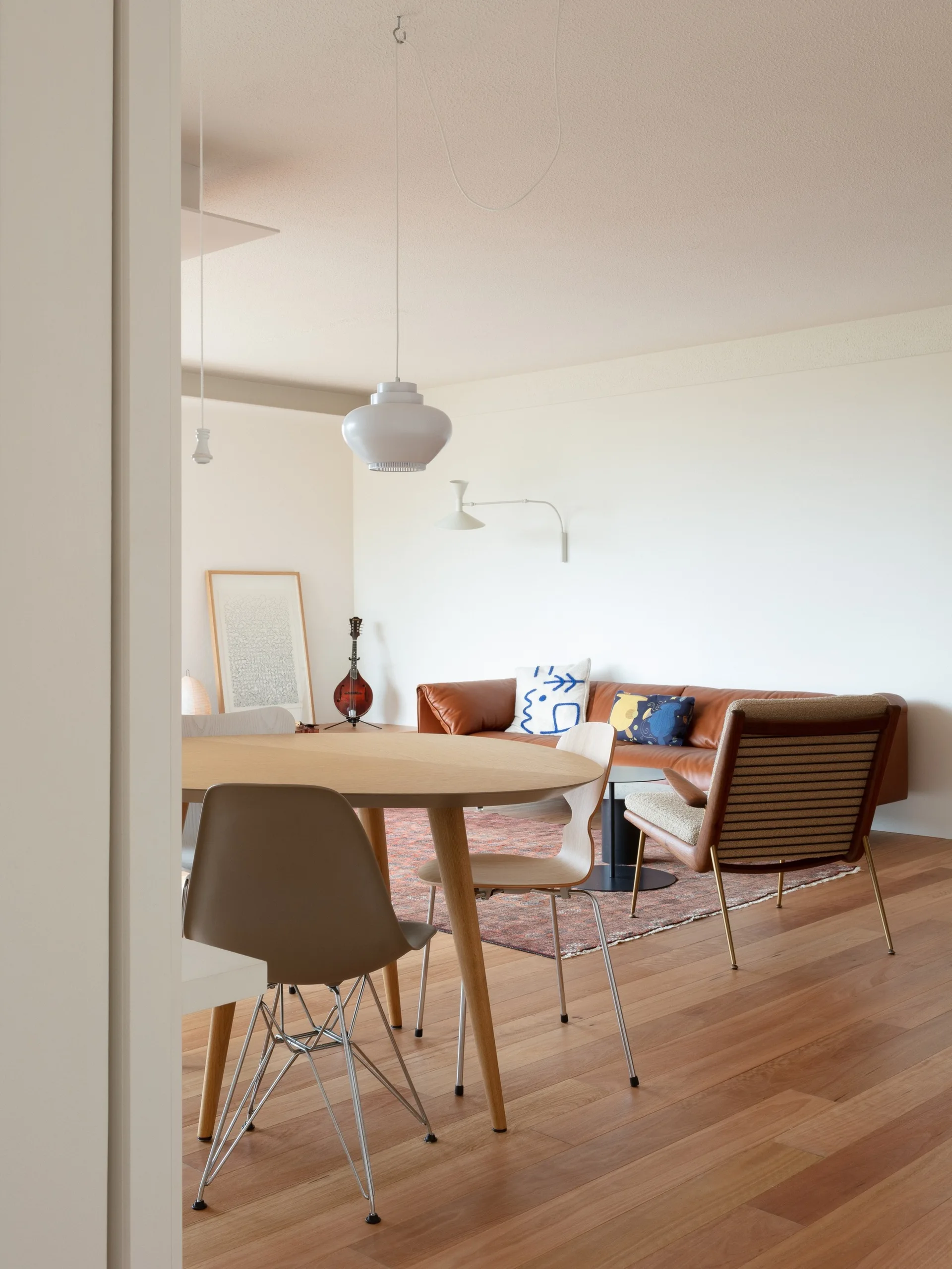 A bright open-plan room features a round wooden dining table with chairs, a brown leather sofa, and a patterned rug on hardwood floors.