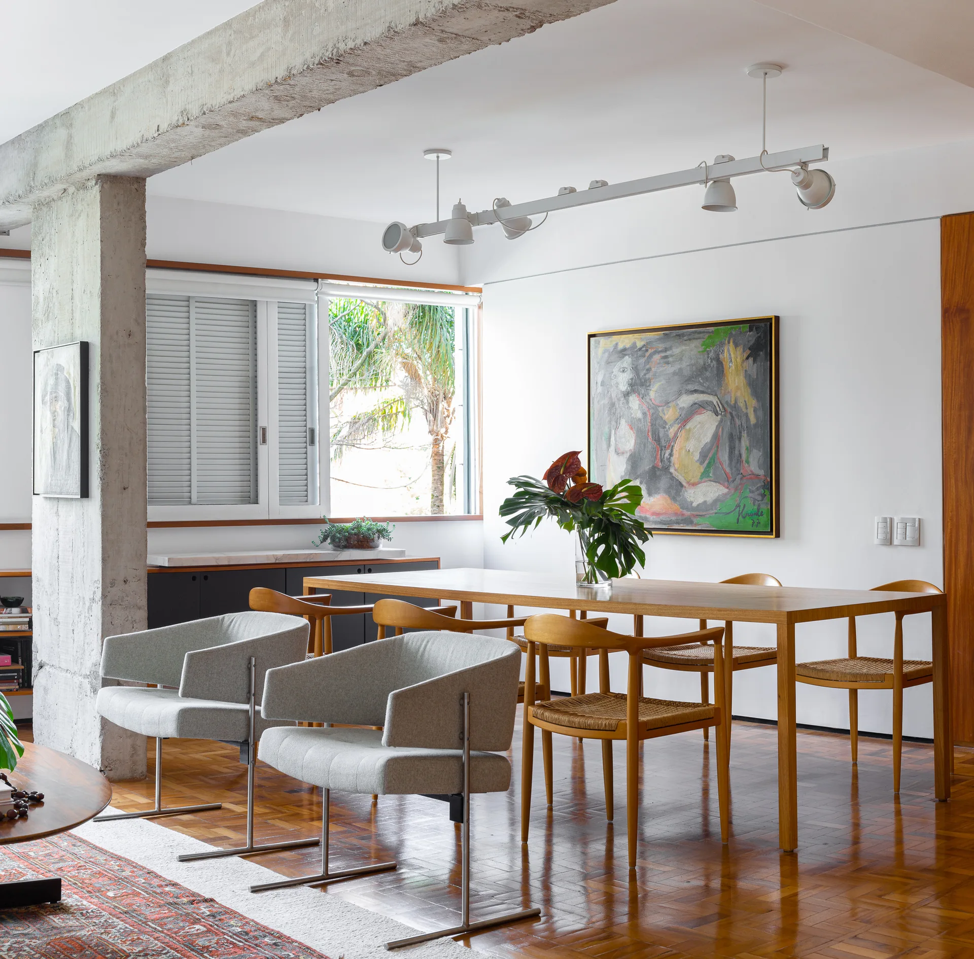 A dining room features a wooden table, mixed wood and grey upholstered chairs, a prominent exposed concrete ceiling beam, and parquet flooring.