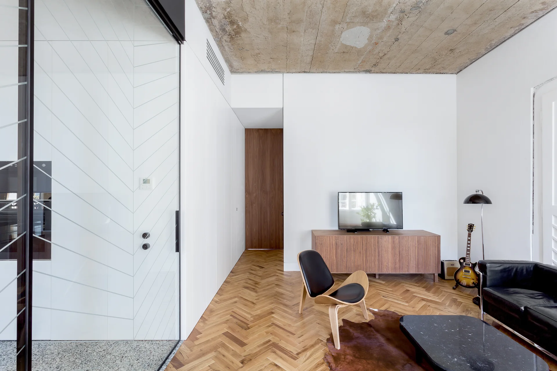 A modern living room features a textured concrete ceiling, herringbone wood floors, a mid-century lounge chair, and a low wooden media console.