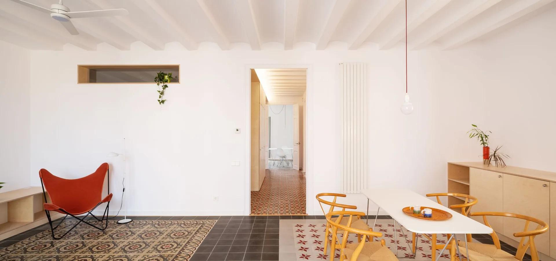 A minimalist living and dining room featuring white walls, exposed ceiling beams, an orange butterfly chair, and decorative patterned floor tiles.