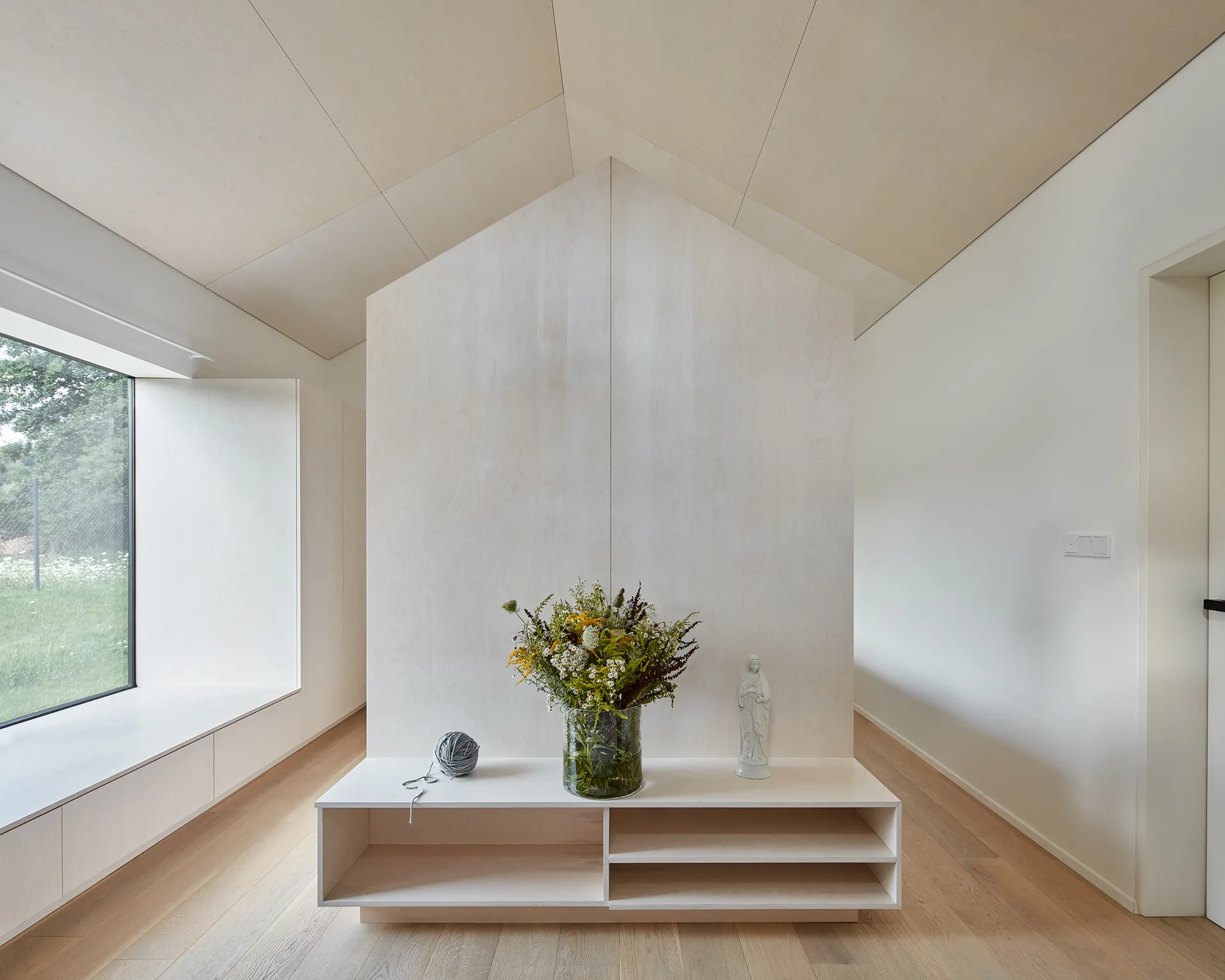 A minimalist room featuring a central house-shaped plywood partition, a low wooden console with flowers, and matching gabled ceiling panels.