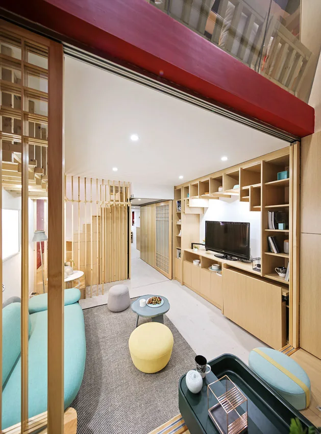 A compact modern living room features a teal sofa, built-in wooden shelving with a TV, and a slatted wood partition.