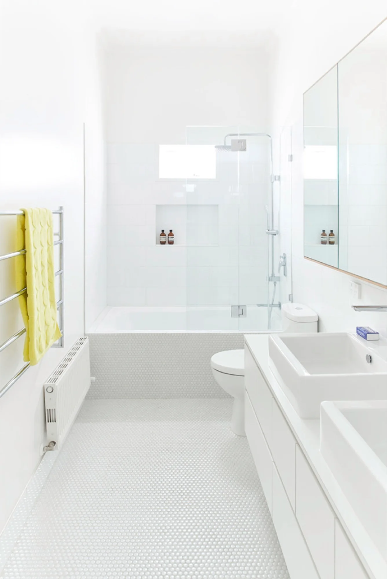 A minimalist white bathroom featuring a double vanity, a recessed bathtub with a glass screen, white penny tile floors, and a bright yellow towel.