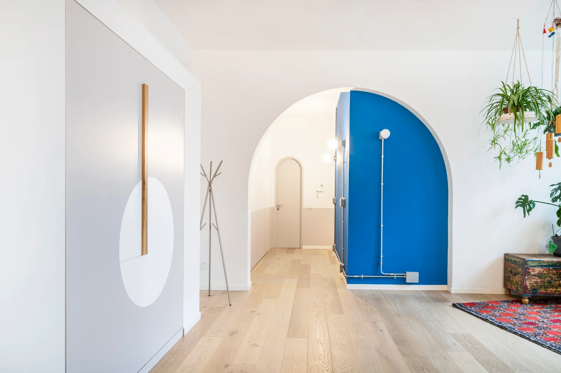A minimalist hallway features a white arched opening looking toward a bright blue accent wall, a grey built-in cabinet, and light wood flooring.