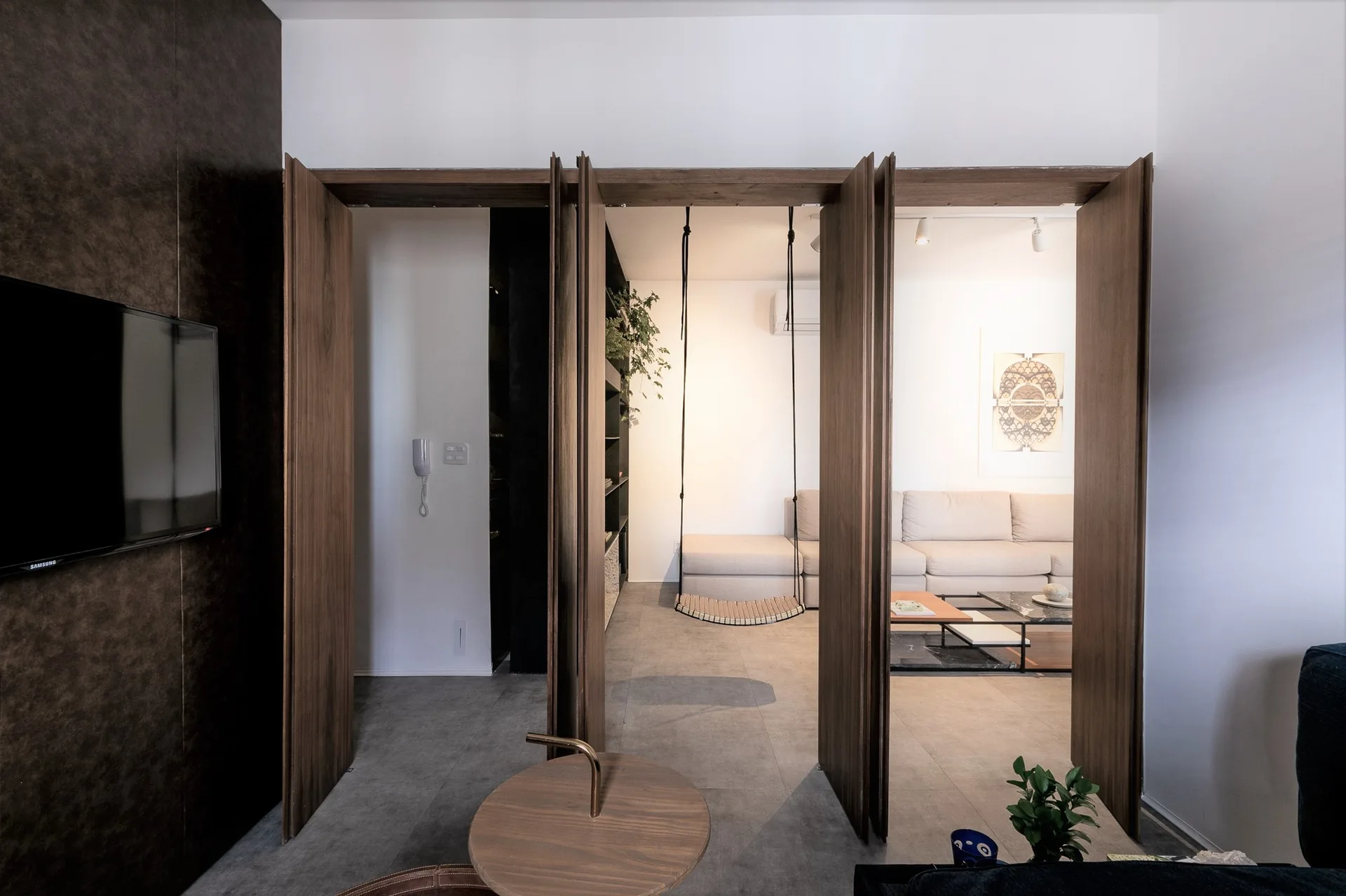Segmented wood folding doors open to reveal an indoor swing chair and a beige sofa in a minimalist living space with grey floor tiles.
