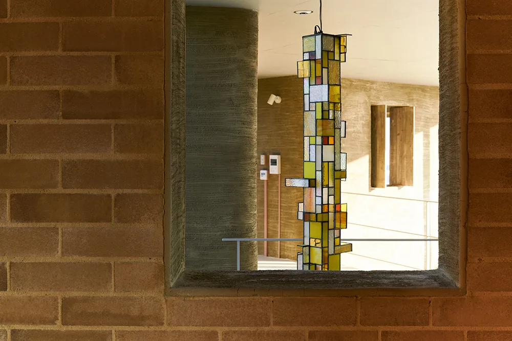 A tall, geometric stained glass pendant light hanging inside a warm concrete space, viewed through a rectangular opening in a brown brick wall.