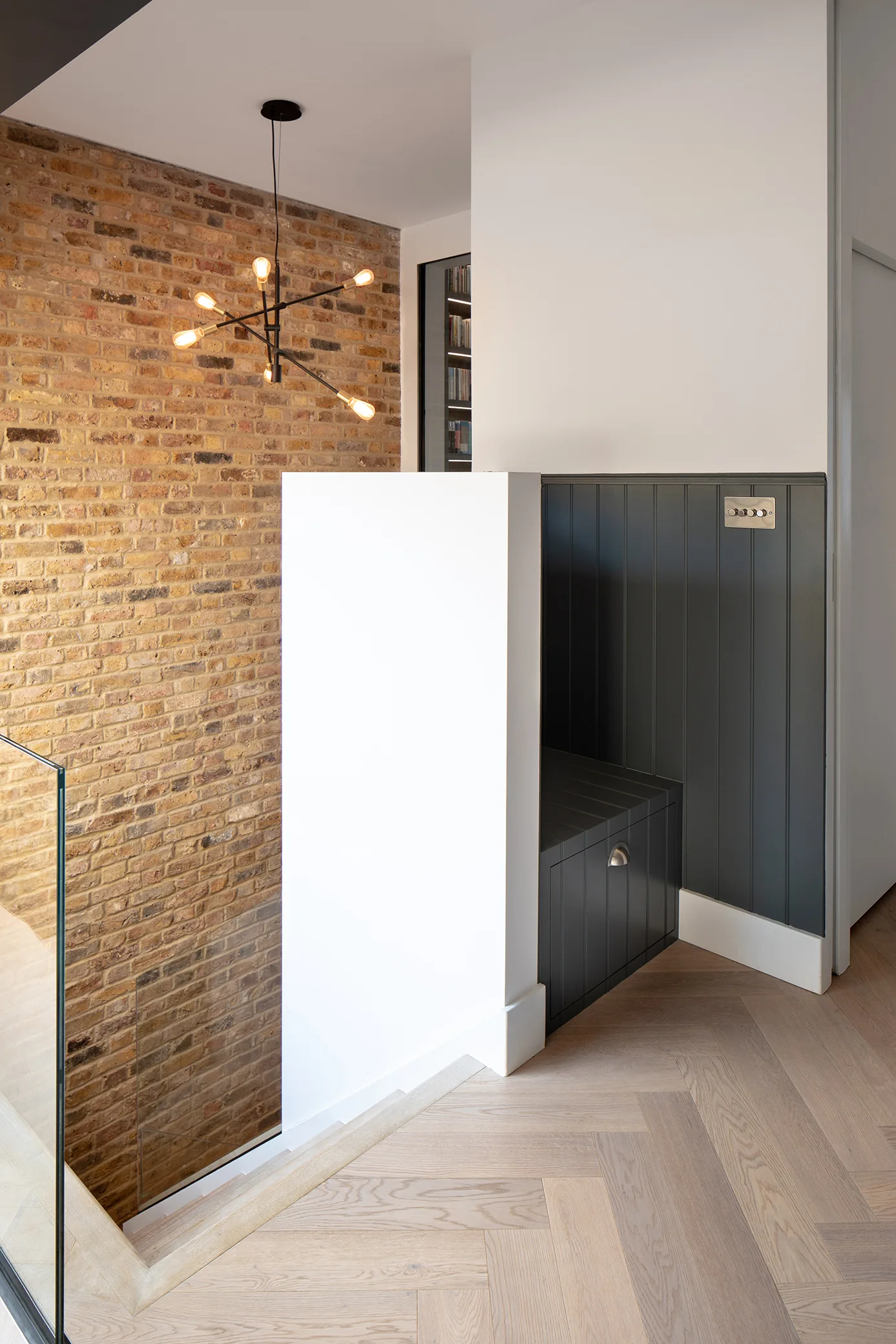 A staircase landing features light herringbone wood floors, a tall exposed brick wall, and dark vertical wood paneling with a built-in bench.