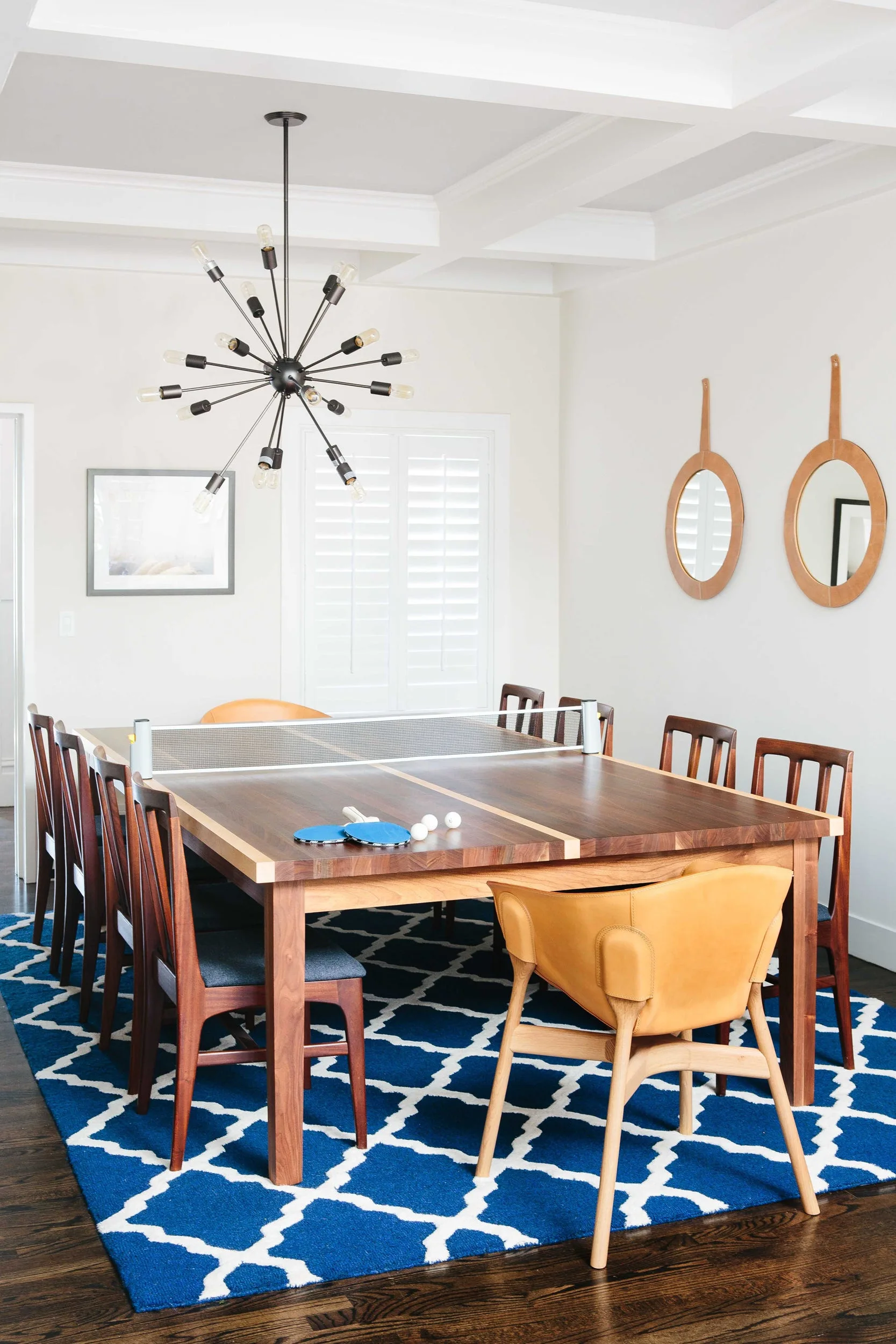 A wooden dining table set up with a ping-pong net sits on a blue patterned rug under a sputnik chandelier in a bright room.