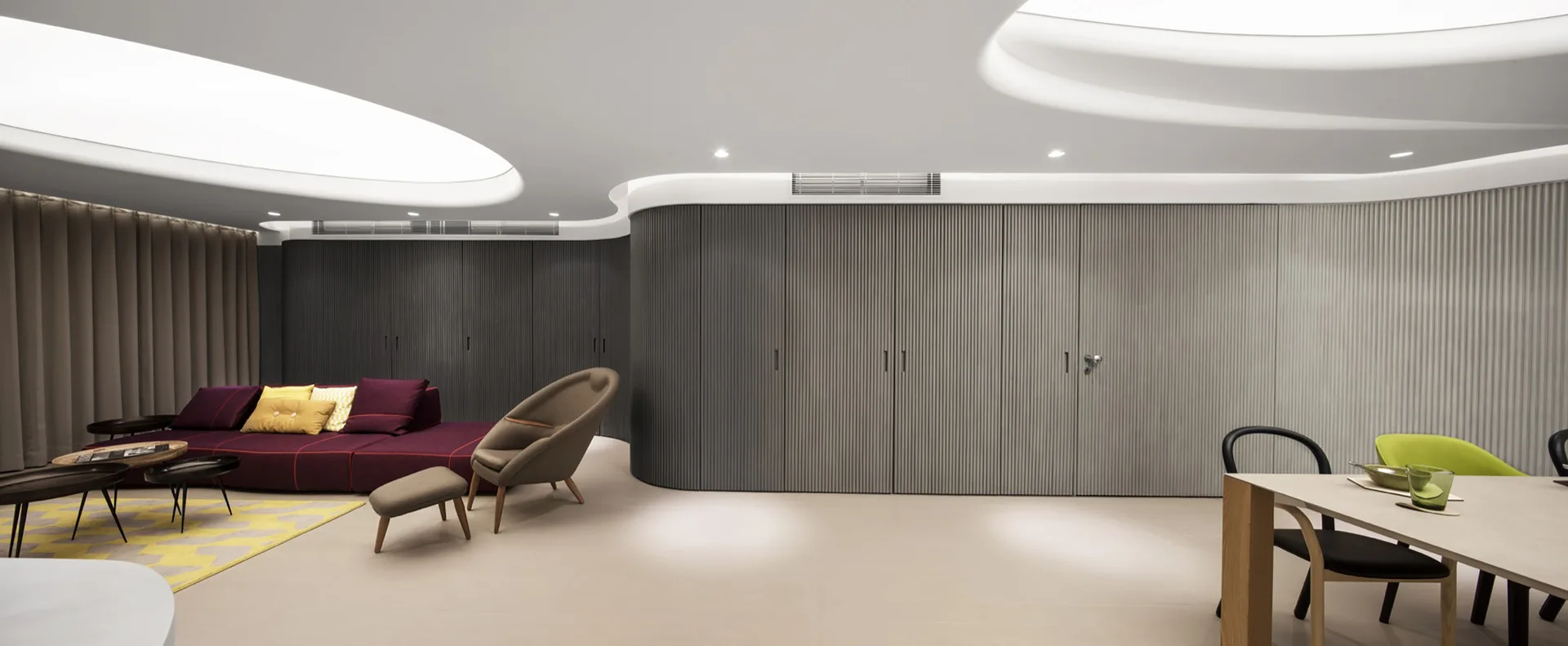 A modern open-plan living space features a large curved wall of grey fluted paneling, a low plum modular sofa, and soft organic ceiling cove lighting.